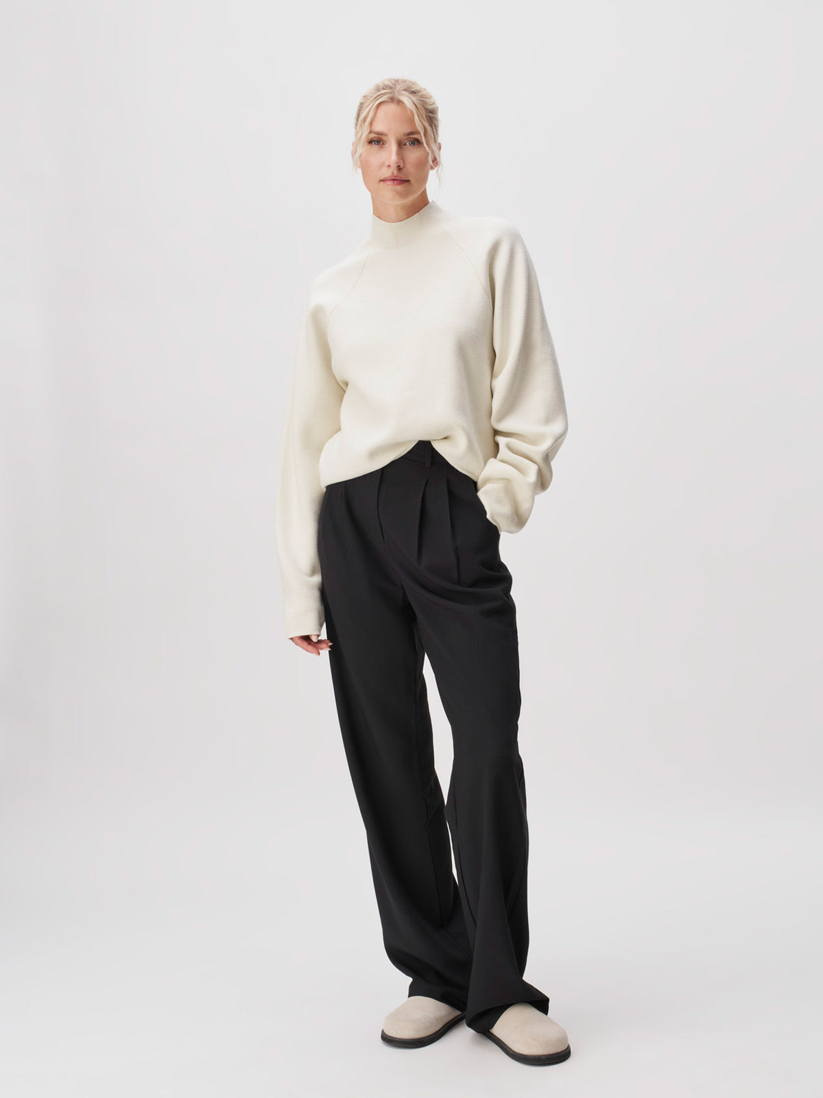 Eine Person steht vor einem schlichten Hintergrund und trägt den Pullover „Heather“ von LeGer by Lena Gercke – einen locker sitzenden weißen Pullover mit langen Ärmeln und normaler Länge –, kombiniert mit einer schwarzen Plisseehose und hellen Slippern. Die Hände hält sie entspannt an den Seiten, ihr Gesichtsausdruck ist neutral.