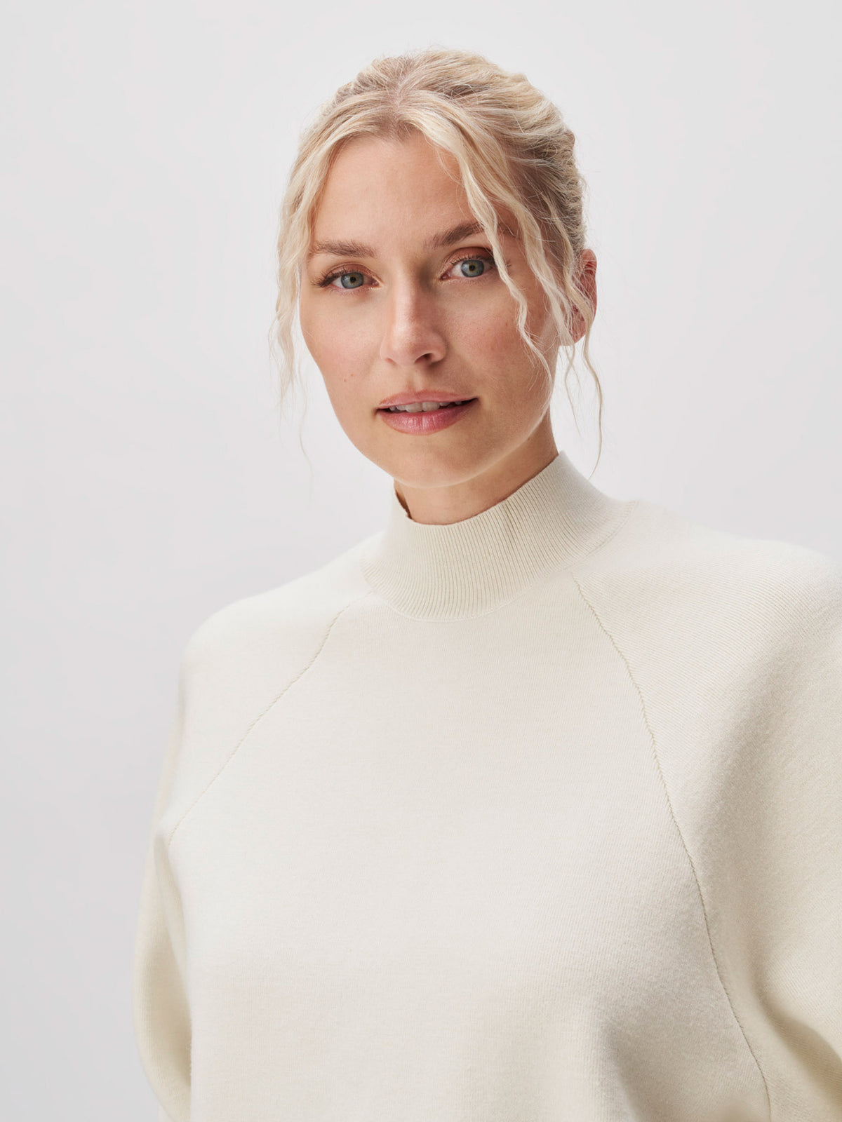 Eine Frau mit blondem, locker gewelltem Haar trägt den cremefarbenen Pullover „Heather“ von LeGer by Lena Gercke, der sich durch lange Ärmel, einen hohen Kragen sowie eine normale Länge und Passform auszeichnet. Sie steht vor einem schlichten, hellen Hintergrund und blickt leicht an der Kamera vorbei.