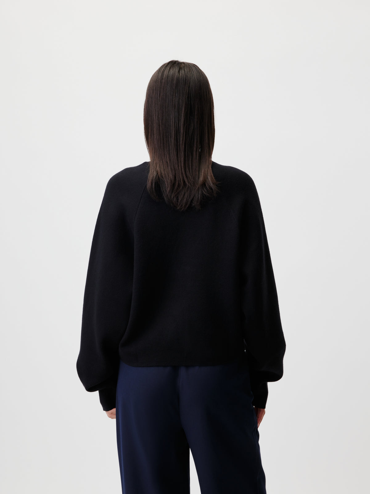 Eine Person mit glattem, schulterlangem, dunklem Haar steht mit dem Rücken zur Kamera und trägt den Pullover „Heather“ von LeGer by Lena Gercke – einen schwarzen Pullover mit Raglanärmeln –, kombiniert mit einer dunklen Hose, vor einem schlichten, hellen Hintergrund.