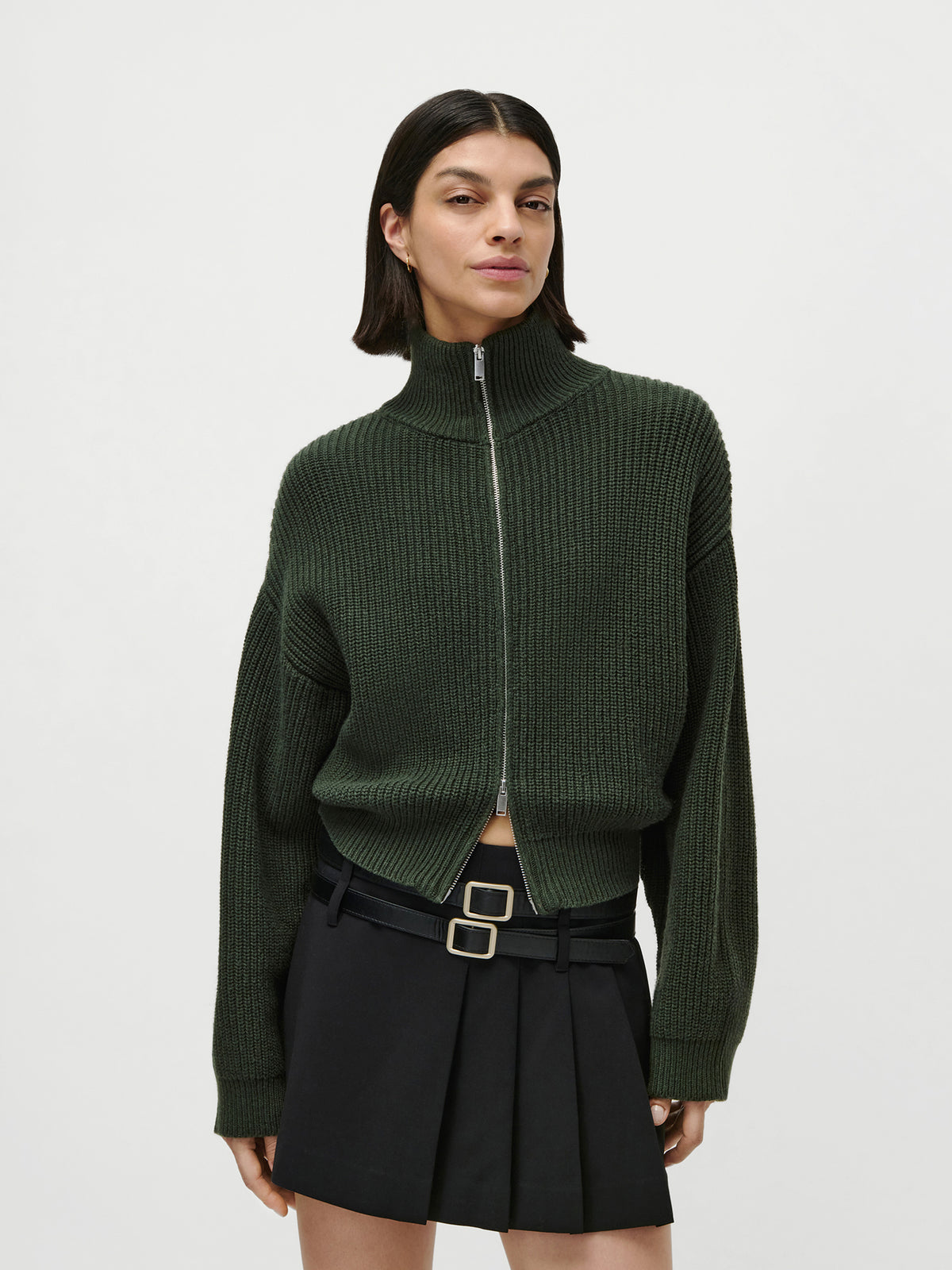 Eine Person, die den Pullover „Cindy“ von LeGer by Lena Gercke trägt – einen dunkelgrünen, gerippten Pullover mit Reißverschluss und langen Ärmeln –, kombiniert mit einem schwarzen, plissierten Minirock mit Gürtel in Größe 36, vor einem schlichten weißen Hintergrund.