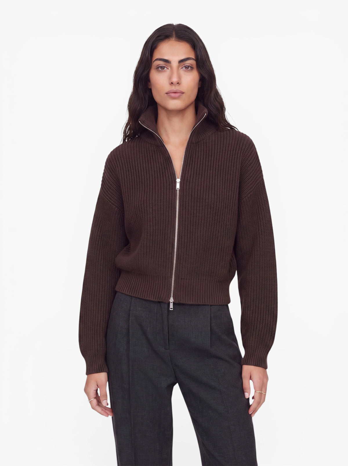 Eine Person mit langen, welligen dunklen Haaren trägt den Pullover „Cindy“ von LeGer by Lena Gercke in Dunkelbraun und eine dunkelgraue Hose und steht vor einem schlichten weißen Hintergrund.