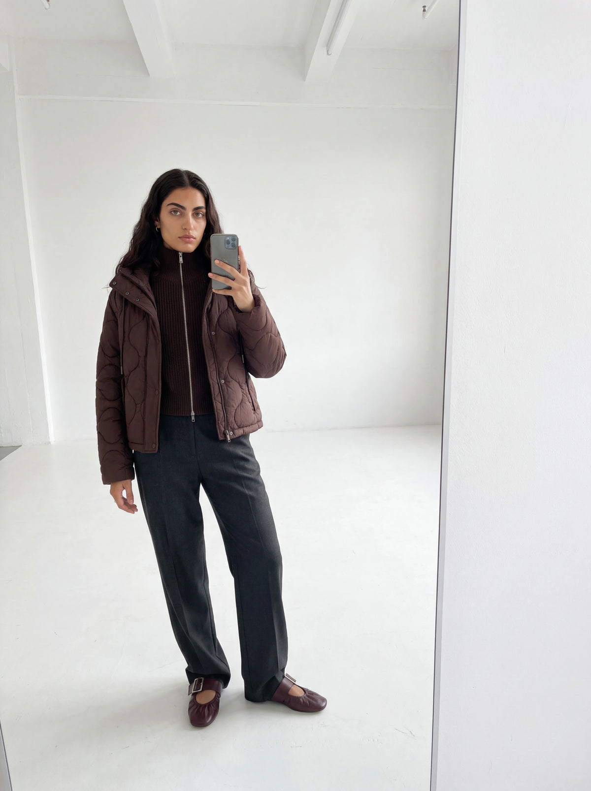 Eine Frau steht in einem minimalistischen, weißen Raum und macht ein Spiegel-Selfie. Sie trägt eine braune Steppjacke über dem „Cindy“-Pullover von LeGer by Lena Gercke – einem dunkelbraunen Rippstrickpullover mit einem dezenten Reißverschlussdetail –, dazu eine dunkle Hose und braune Schuhe. Der Hintergrund ist hell und aufgeräumt.