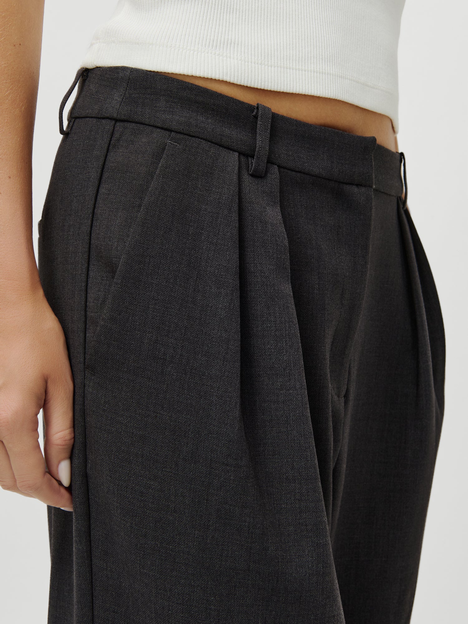 Eine Nahaufnahme zeigt eine Person, die die LeGer x Oh April - Franja Pants Tall von LeGer by Lena Gercke in Dunkelgrau mit hoher Taille und Bundfalten trägt, gepaart mit einem weißen gerippten Tanktop; die Passform passt zu denen, die Größe 38 suchen. Die Hand ruht lässig an der Seite.