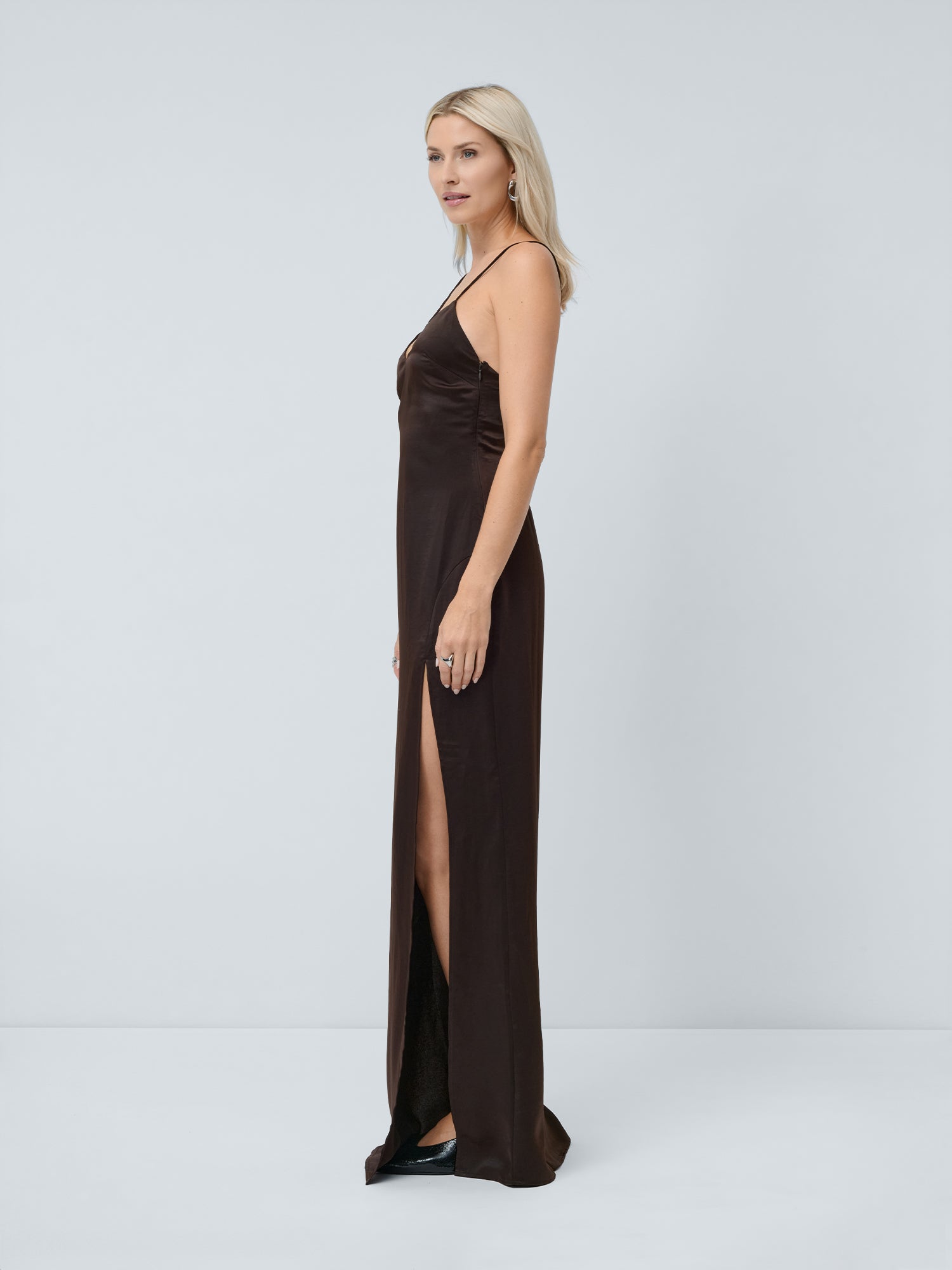 Eine Frau mit hellem Haar trägt das „Lena“ Slim-Fit-Kleid von LeGer by Lena Gercke mit Spaghettiträgern und einem oberschenkellangen Schlitz und steht seitlich vor einem schlichten hellgrauen Hintergrund.