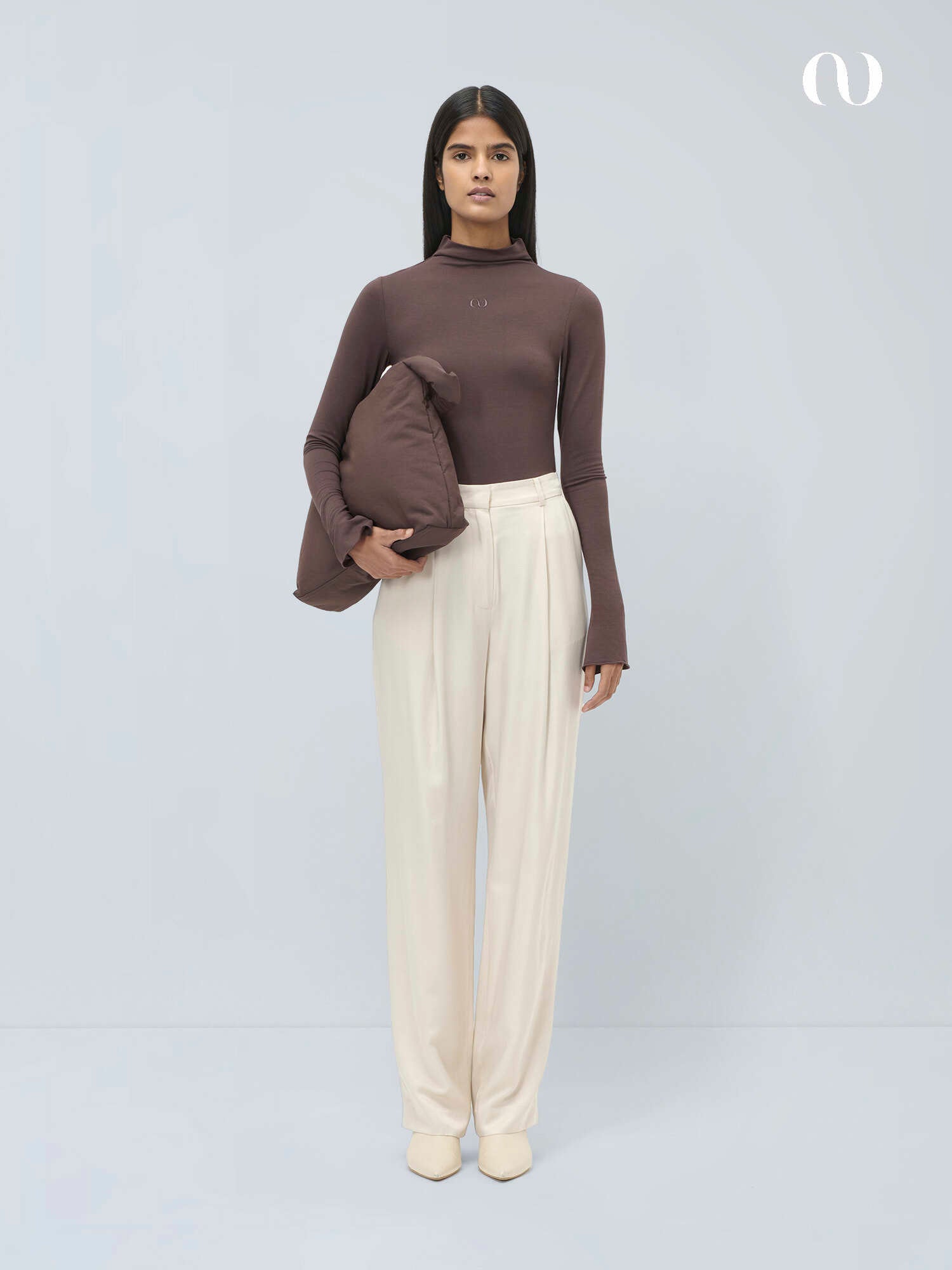 Eine Frau mit glattem dunklem Haar trägt den braunen LeGer by Lena Gercke Bessy Longsleeve-Rollkragenpullover und eine cremefarbene Hose mit hoher Taille. Sie hält eine passende braune wattierte Jacke unter dem Arm vor einem schlichten hellgrauen Hintergrund.