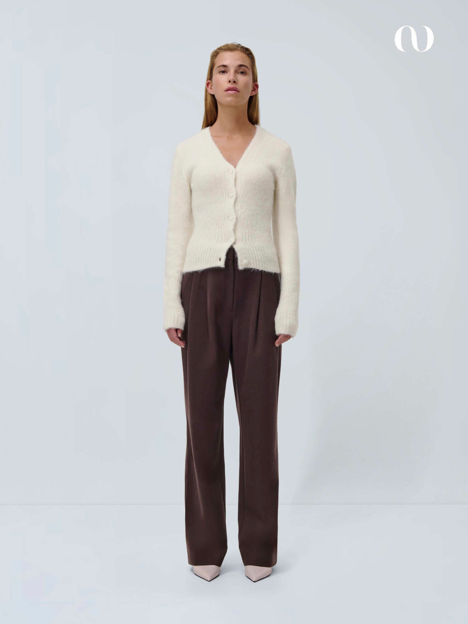 Eine Frau steht vor einem schlichten, hellen Hintergrund und trägt die Inken Pants von LeGer by Lena Gercke in Dunkelbraun (Maxi wide-leg, Größe 36), eine cremefarbene Strickjacke und helle spitze Schuhe. Sie blickt nach vorne und hat einen neutralen Gesichtsausdruck. Ein kleines weißes Logo befindet sich oben rechts.