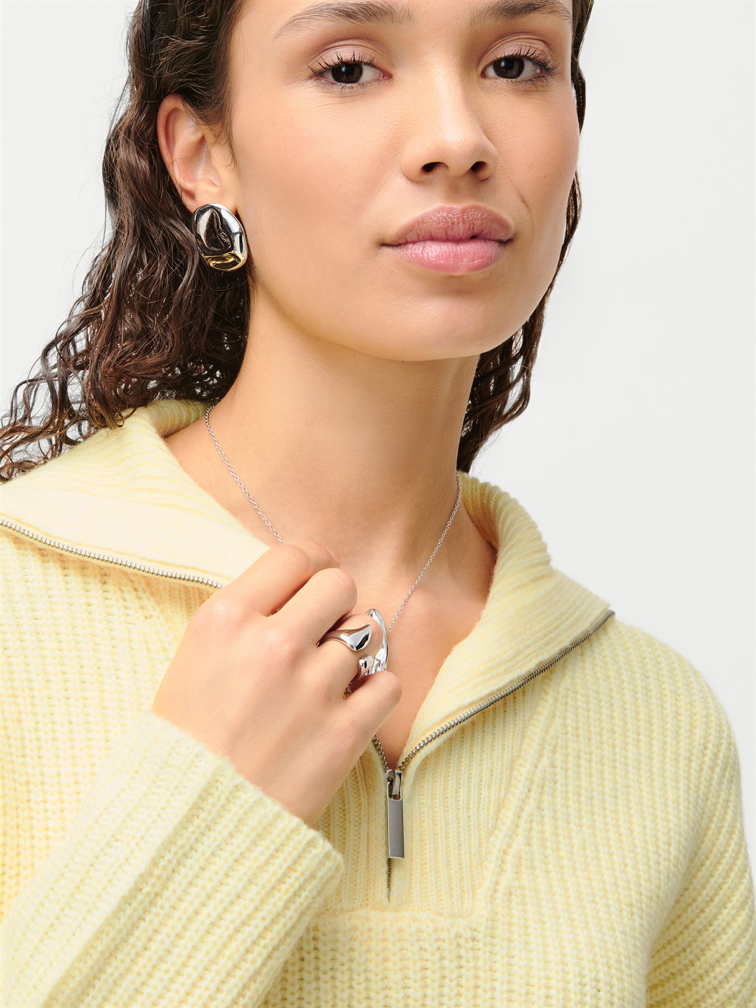 Eine Frau trägt den hellgelben LeGer x Purelei - Lea Pullover von LeGer by Lena Gercke mit einem Reißverschluss, einer lockeren Passform und extra langen Ärmeln. Dazu trägt sie große silberne Ohrringe, einen Ring und eine Halskette. Ihre Hand ruht in der Nähe ihres Kragens.