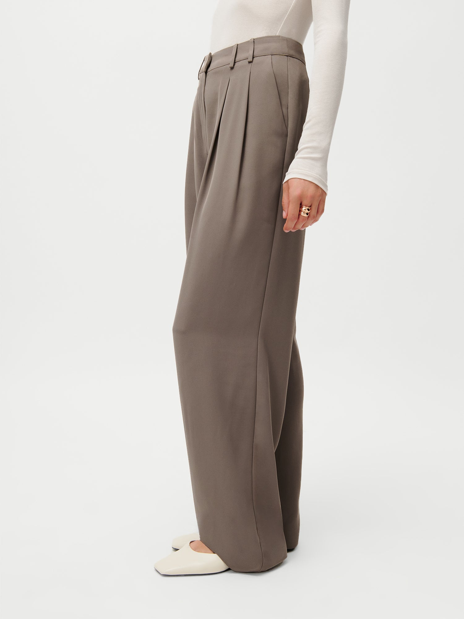 Ein Model (1,72m, Größe 36) trägt die LeGer x Purelei Cara Pants von LeGer by Lena Gercke - eine braune Hose mit weitem Bein und hoher Taille - kombiniert mit einem weißen, langärmeligen Top, cremefarbenen, spitzen Schuhen und goldenen Ringen vor einem schlichten weißen Hintergrund.