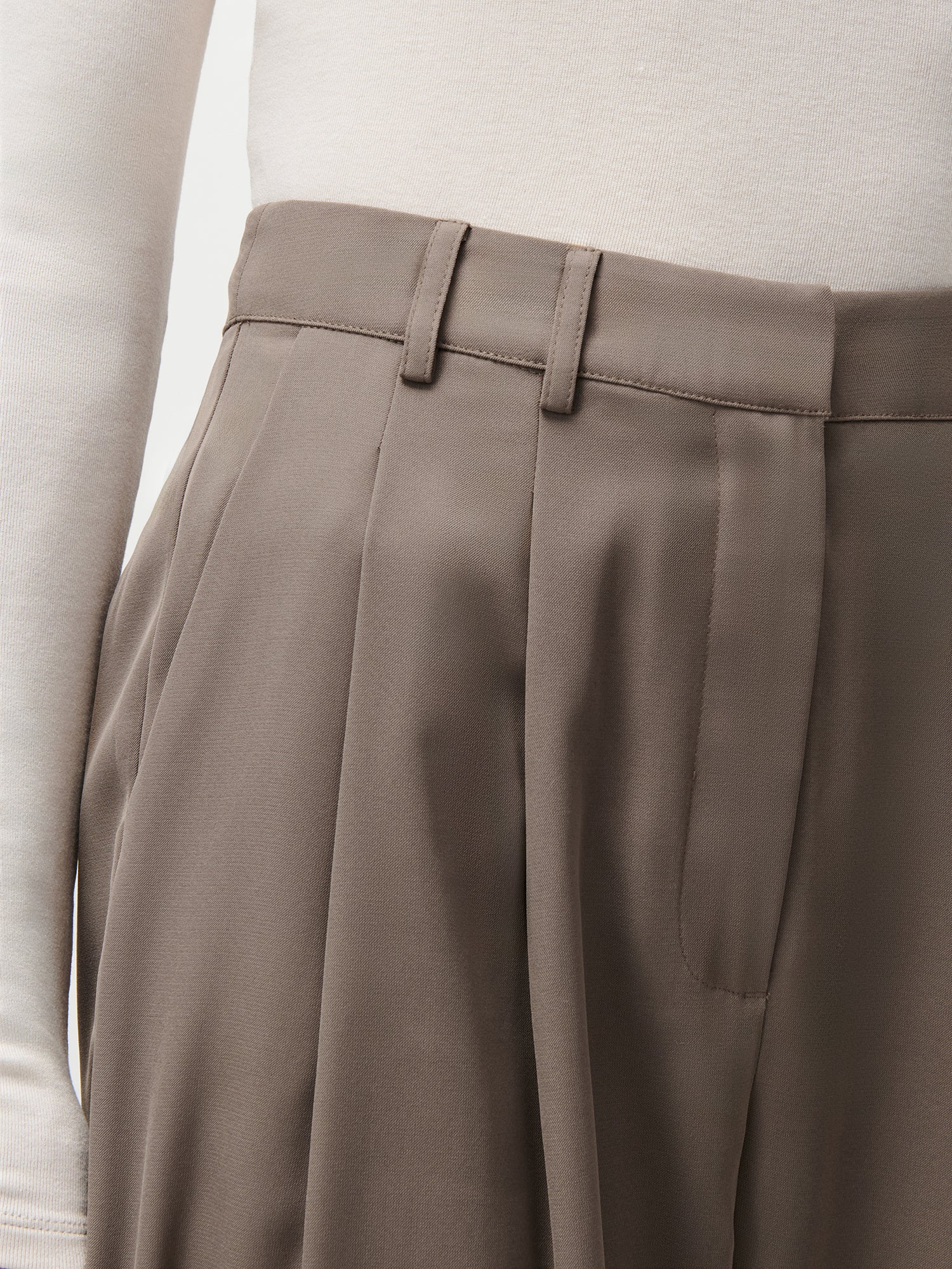 Nahaufnahme einer Person (Größe 36, 1,72m), die die LeGer x Purelei - Cara Pants by LeGer by Lena Gercke trägt: hochtaillierte taupefarbene Bundfaltenhose, kombiniert mit einem taillierten, langärmeligen, cremefarbenen Oberteil; nur Taille und ein Teil des Oberkörpers/Arms sind sichtbar.