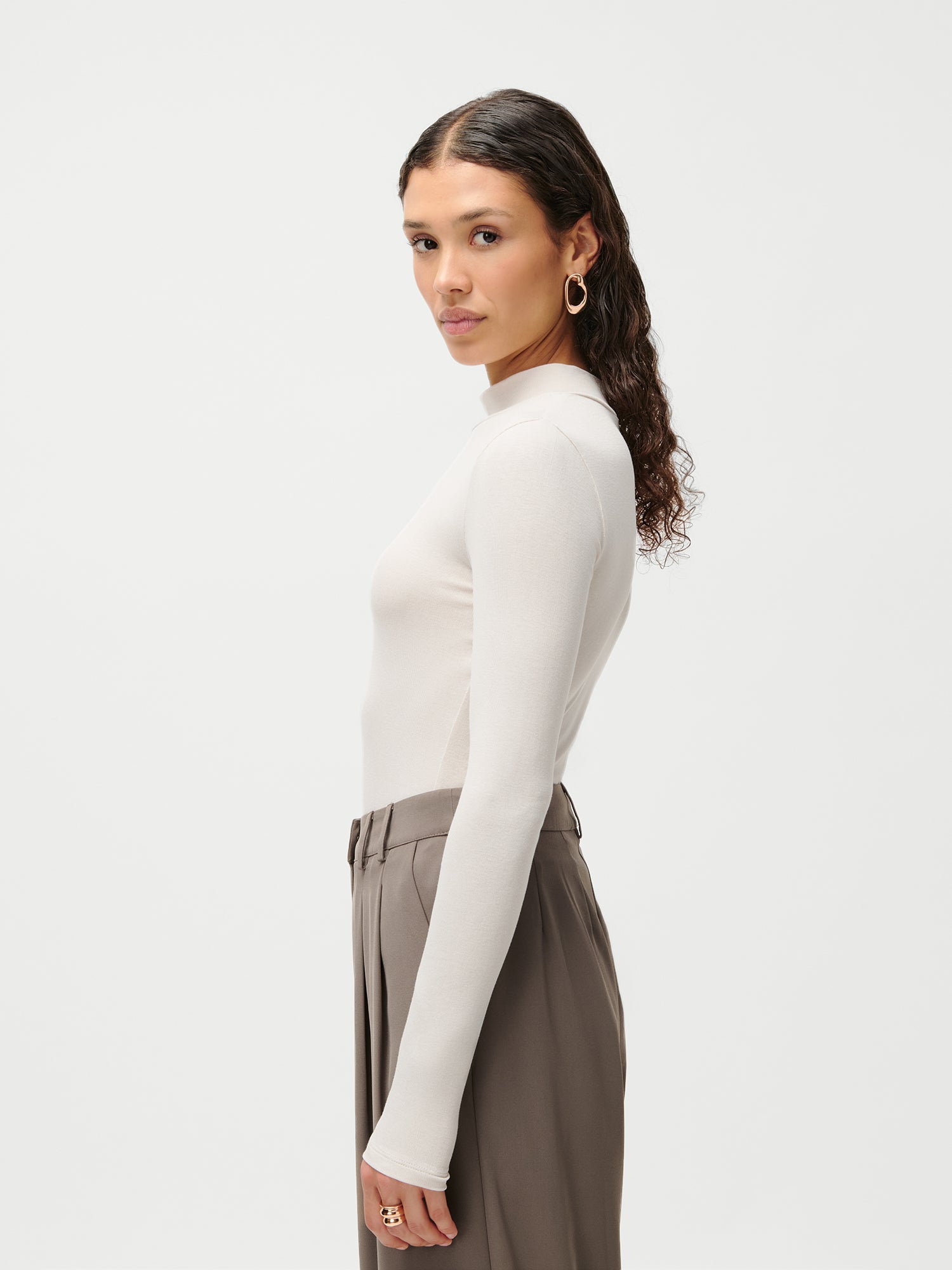 Eine Frau mit gewellten braunen Haaren, die ein LeGer x Purelei - Deike Longsleeve von LeGer by Lena Gercke in Weiß (Größe 36) und eine taupefarbene Hose mit hoher Taille trägt, steht im Profil vor einem einfarbig weißen Hintergrund mit einem neutralen Ausdruck.