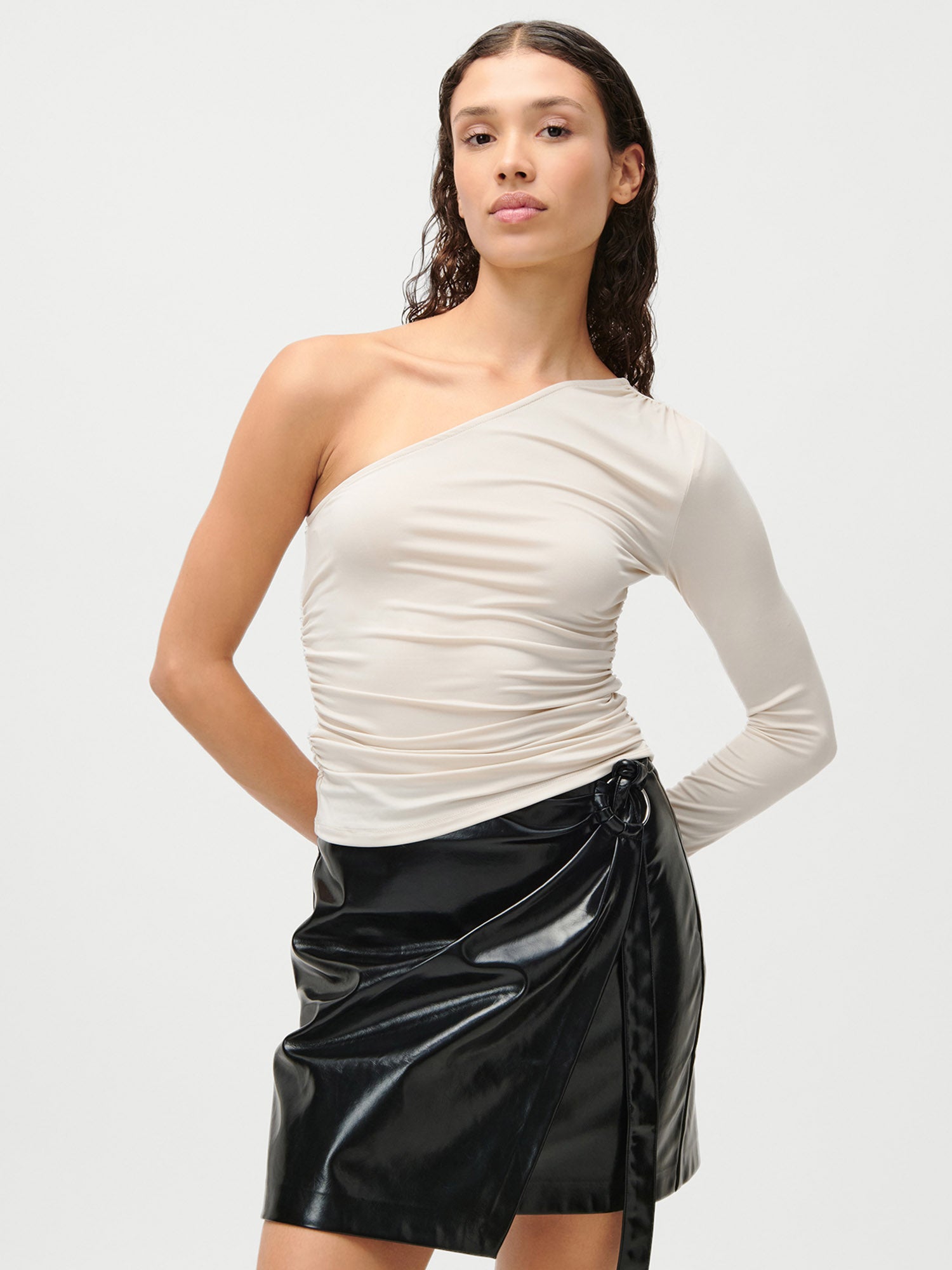 Eine Person mit langen Haaren im Wetlook trägt das beigefarbene Sophia Top von LeGer by Lena Gercke (Slim Fit, langarm) und einen glänzenden schwarzen Wickelrock in Größe 36 und steht selbstbewusst vor einem schlichten hellen Hintergrund.