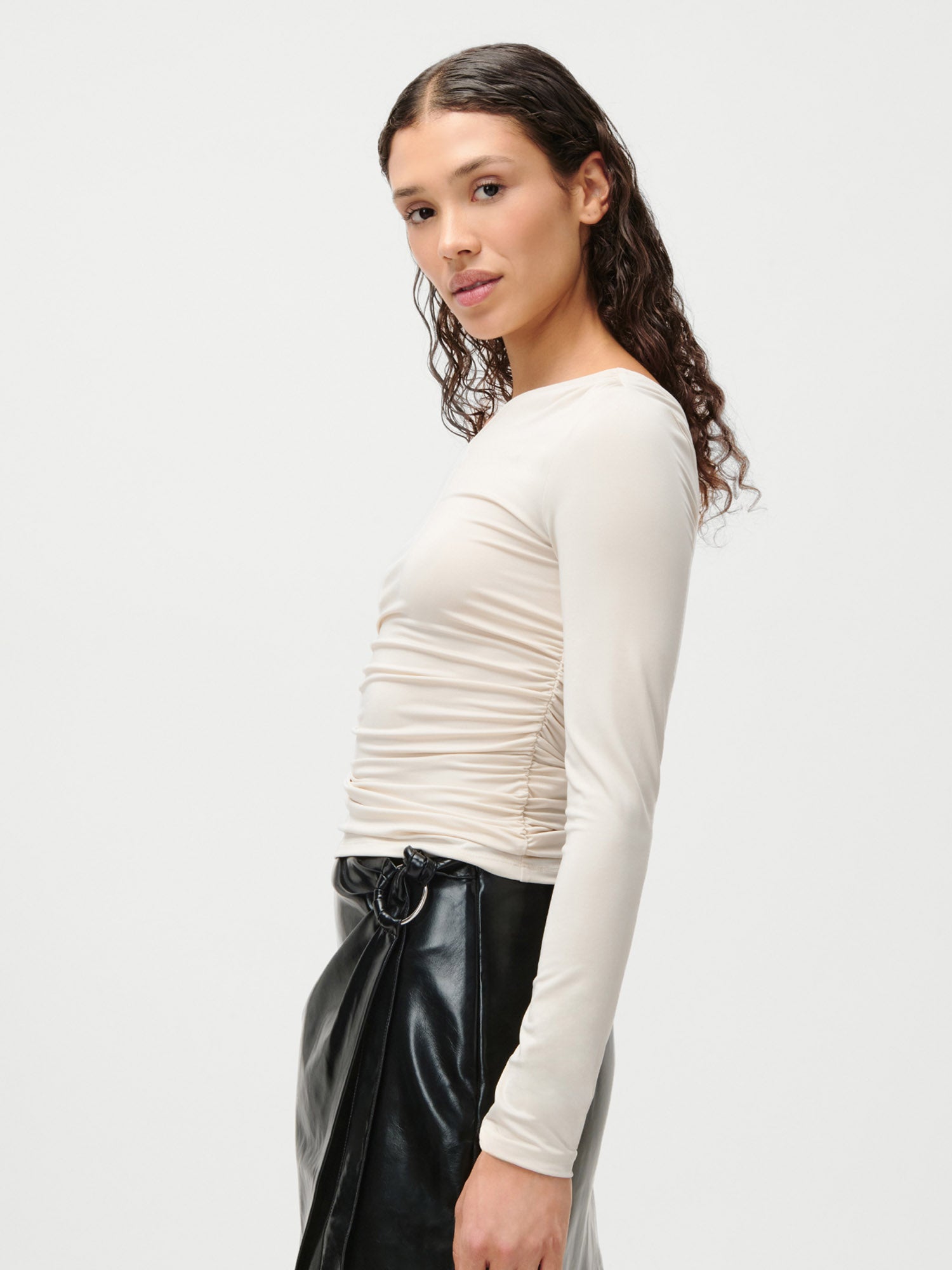 Eine Frau mit langen, gewellten braunen Haaren trägt das LeGer by Lena Gercke Sophia Top in Beige, Größe 36, gestylt mit einem glänzenden schwarzen Rock, im Profil vor einem schlichten weißen Hintergrund.