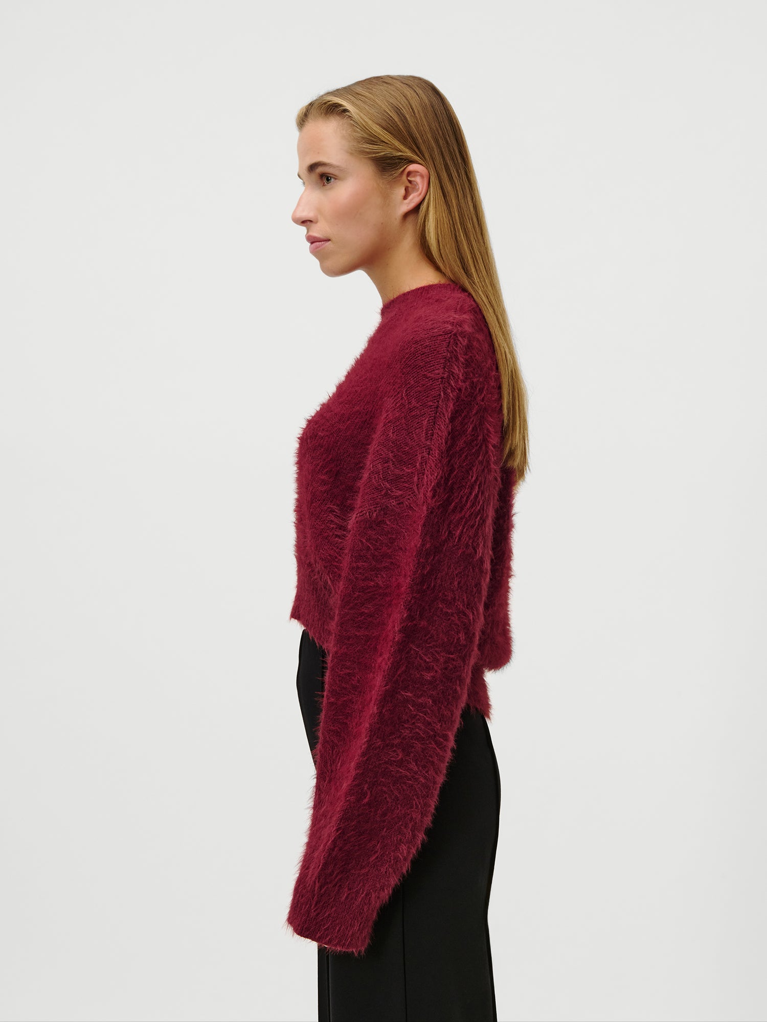 Eine Person mit langem, glattem Haar modelliert den wuscheligen, dunkelroten Celestine-Pullover von LeGer by Lena Gercke und eine schwarze Hose in Größe 36. Sie steht im Profil vor einem schlichten weißen Hintergrund.