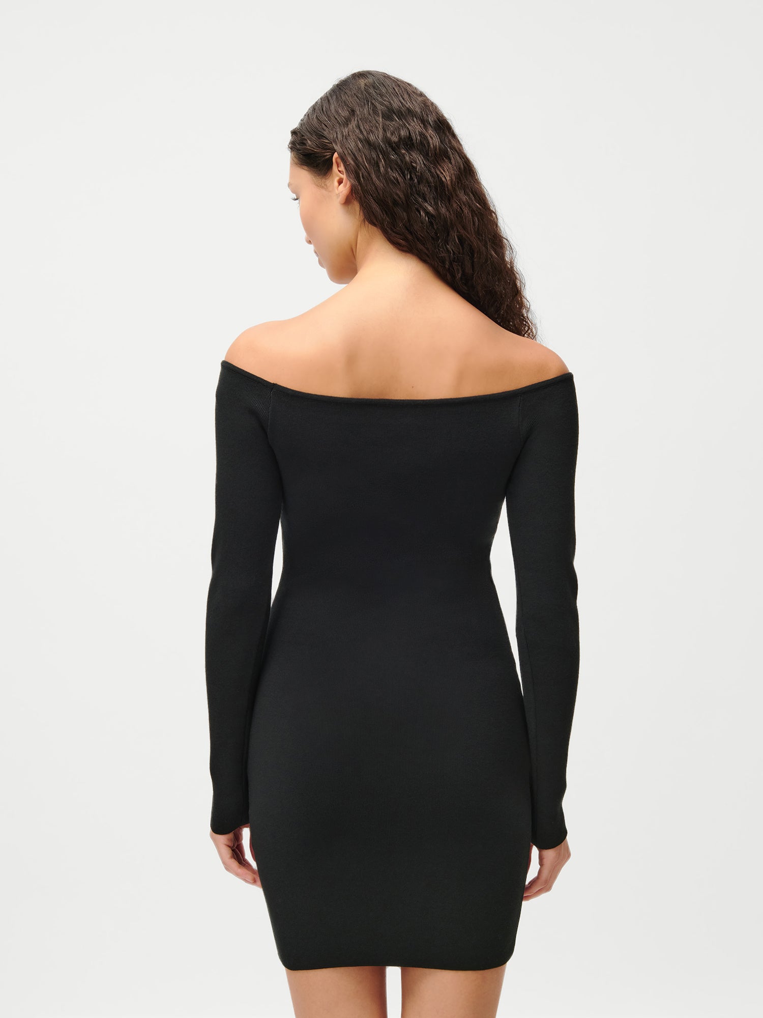 Eine Frau mit lockigem braunem Haar modelliert das Evelin-Kleid von LeGer by Lena Gercke - ein schmal geschnittenes, schulterfreies schwarzes Kleid mit langen Ärmeln. Sie steht mit dem Rücken zur Kamera vor einem schlichten weißen Hintergrund.