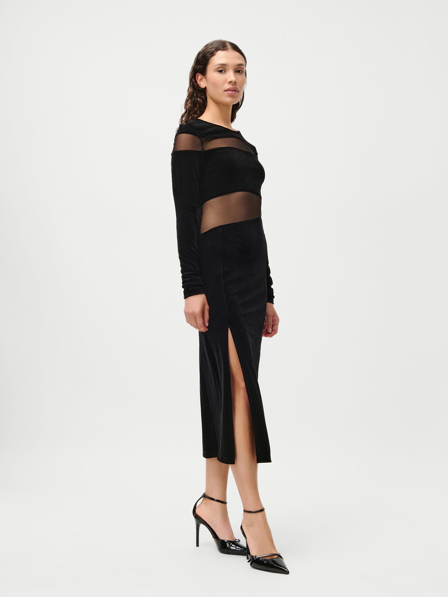 Eine Frau trägt das LeGer by Lena Gercke Fray Dress - ein schwarzes, 7/8 langes, langärmeliges Kleid mit durchsichtigen Mesh-Einsätzen und einem oberschenkelhohen Schlitz - kombiniert mit schwarzen Absätzen vor einem schlichten weißen Hintergrund.