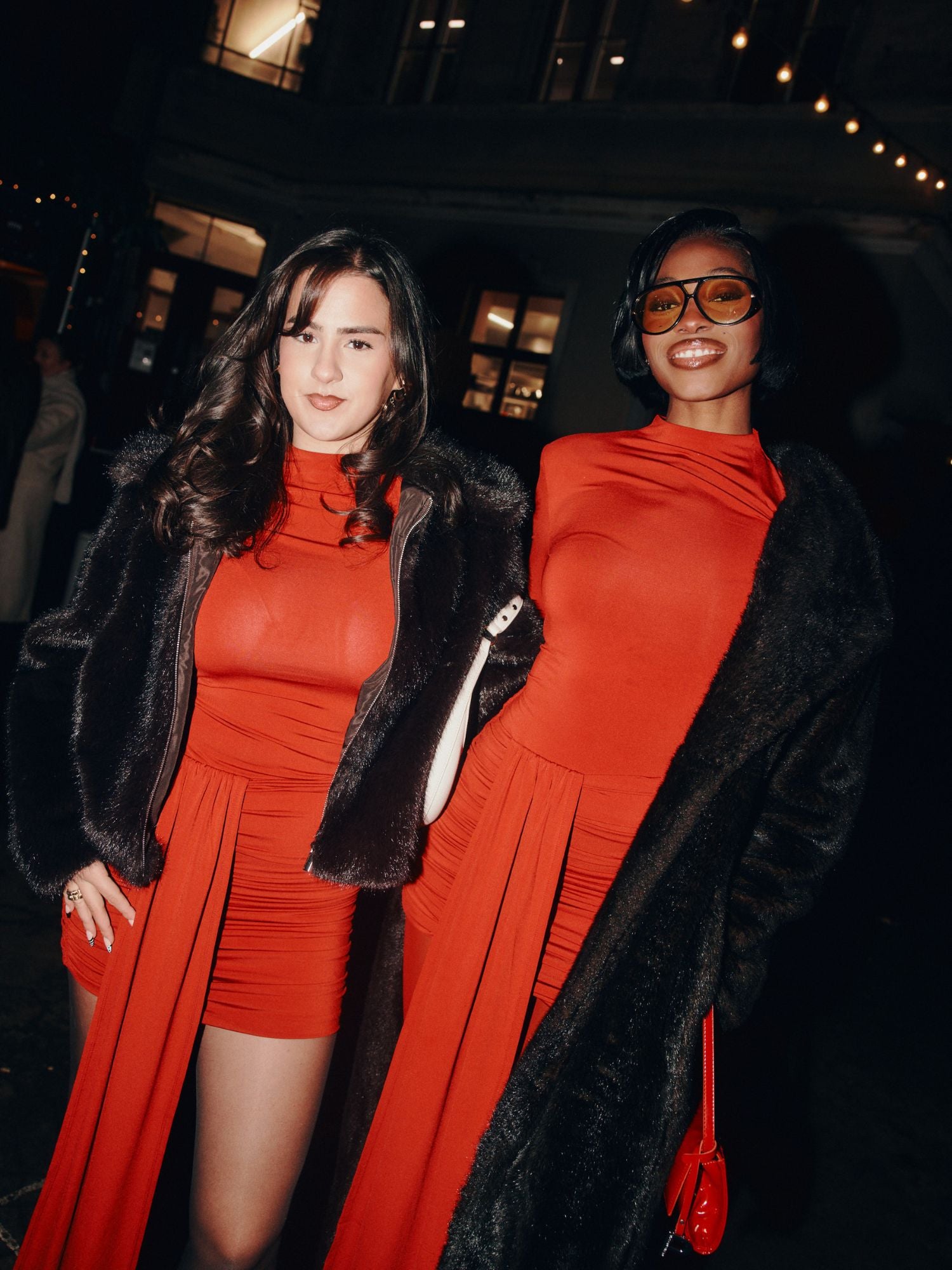 Zwei Frauen stehen nachts im Freien, beide lächeln und posieren für die Kamera. Sie tragen passende rote langärmelige Kleider in Größe 36 und schwarze Catherine Jacken von LeGer by Lena Gercke. Die eine hat langes braunes Haar, die andere hat kurzes Haar und eine große Brille.