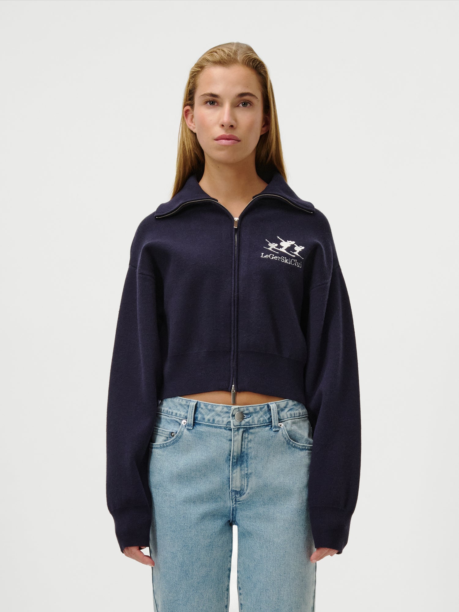 Eine junge Frau mit langem, glattem blondem Haar trägt den LeGer by Lena Gercke Jaden Jumper, eine marineblaue Kurzjacke mit Reißverschluss und extralangen Ärmeln, und eine hellblaue High-Waisted-Jeans in Größe 36 vor einem schlichten weißen Hintergrund.