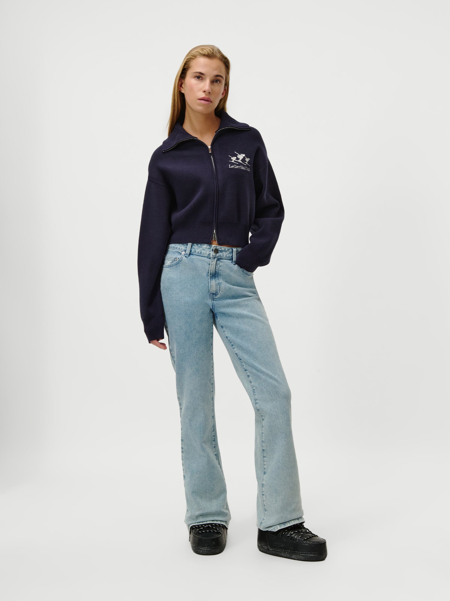 Eine Frau steht vor einem weißen Hintergrund und trägt den Jaden-Pullover von LeGer by Lena Gercke - marineblau mit extralangen Ärmeln und einem kleinen weißen Logo - in Kombination mit einer hellen Jeans in Größe 36 und schwarzen Plateauschuhen, die linke Hand in der Tasche.