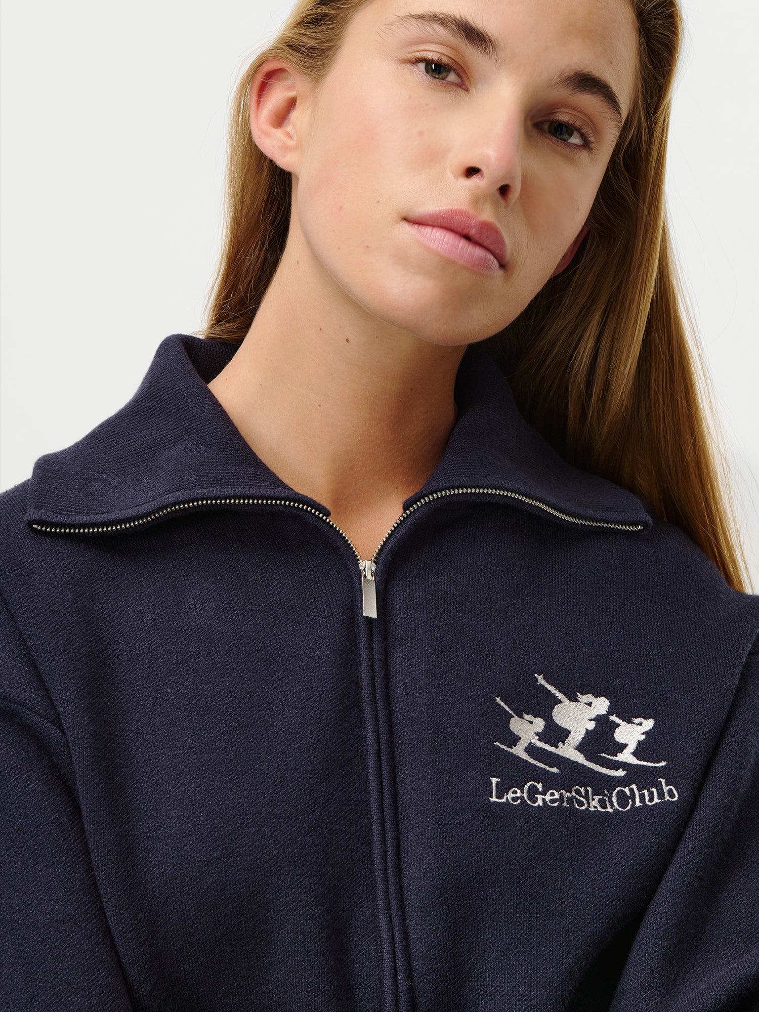 Eine Frau mit langen hellbraunen Haaren trägt den LeGer by Lena Gercke Jaden Jumper in Größe 36, einen marineblauen Pullover mit extra langen Ärmeln und einem "LeGer Ski Club" Logo mit drei weißen Skifahrern auf der Brust. Sie blickt mit einem neutralen Blick nach oben.