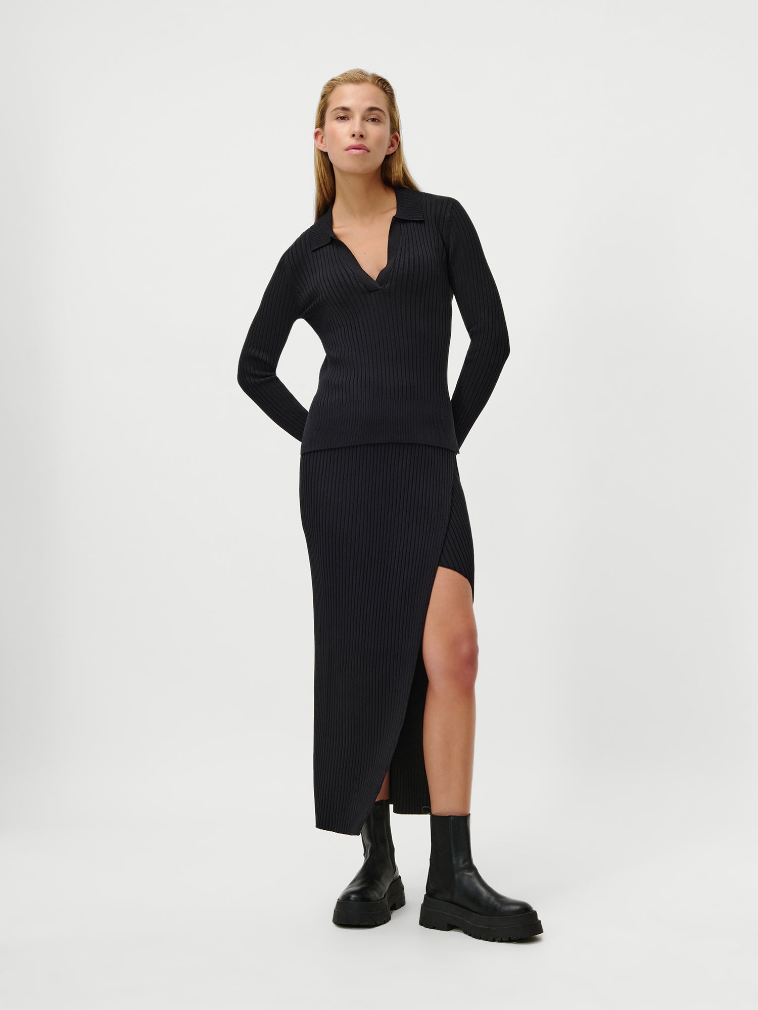 Eine Frau posiert vor einem schlichten Hintergrund mit dem LeGer by Lena Gercke Charlotte Jumper, einem taillierten schwarzen Ripp-Top mit extralangen Ärmeln und V-Ausschnitt, zu dem ein passender Midi-Rock mit hohem Schlitz und klobige schwarze Stiefel getragen werden.