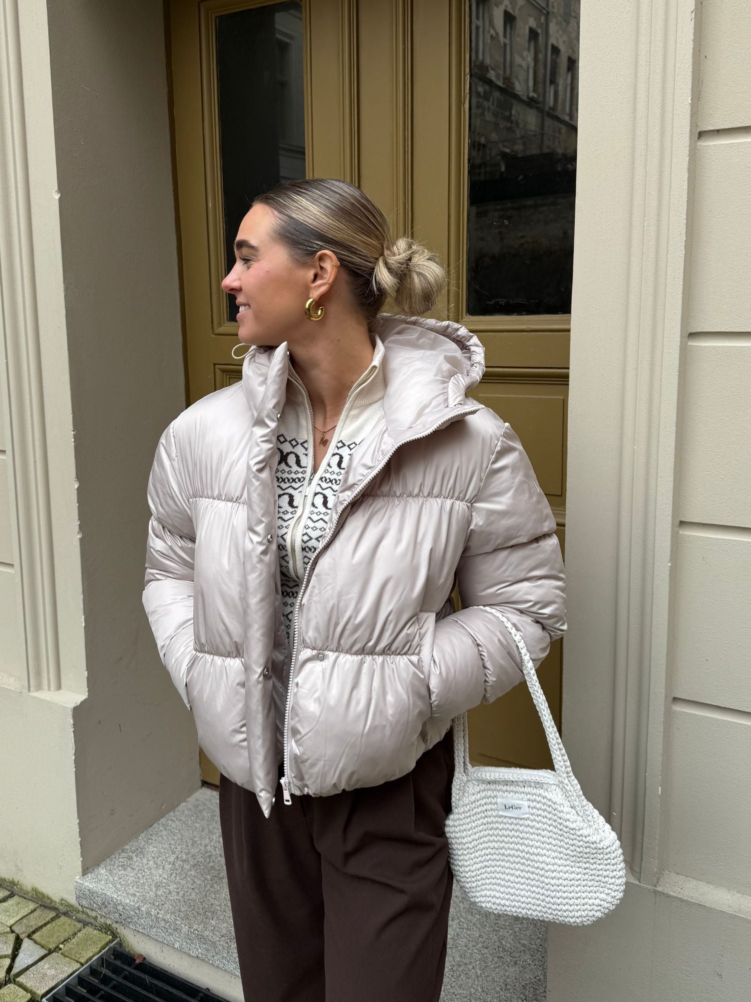 Eine blonde Frau mit Dutt steht im Freien und trägt die LeGer by Lena Gercke Viveka Jacke mit langen Ärmeln, eine braune Hose und goldene Ohrringe. Sie hält eine weiße, strukturierte Handtasche in der Hand und lächelt sanft. Geeignet für Größe 36.