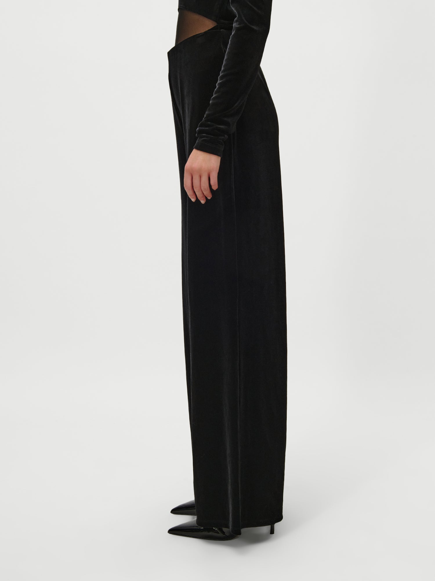Eine Person steht seitlich in der schwarzen Samthose „Fray“ von LeGer by Lena Gercke – mit hoher Taille, weitem Bein und Maxilänge – und trägt dazu ein langärmeliges schwarzes Oberteil sowie spitze schwarze Pumps vor einem schlichten weißen Hintergrund.