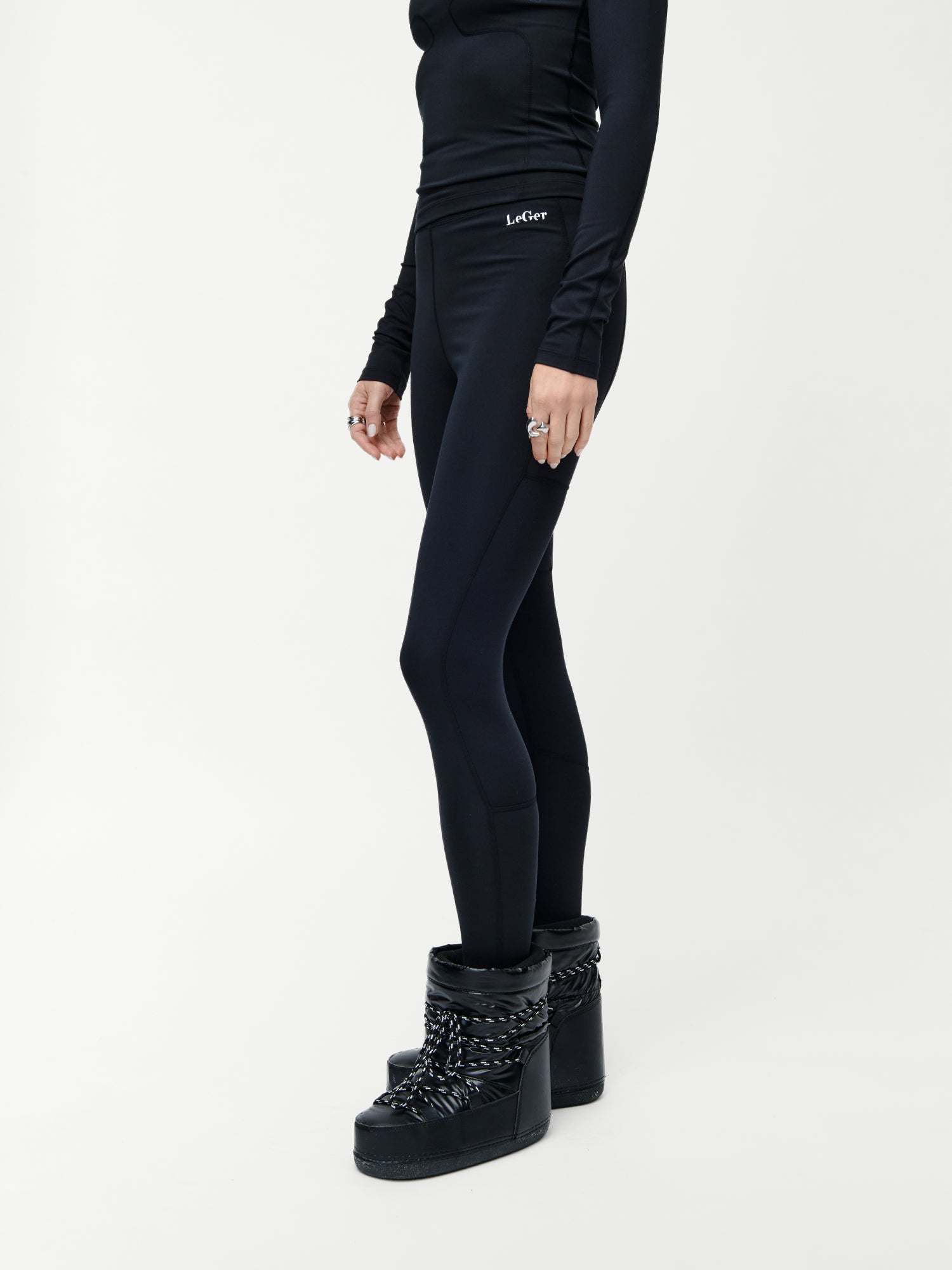 Eine Person, die eine LeGer by Lena Gercke Thalke Hose, ein schwarzes langärmeliges sportliches Oberteil und Maxi Plateau-Winterstiefel mit Kettendetails trägt, steht vor einem schlichten weißen Hintergrund. Das Gesicht der Person ist nicht sichtbar.