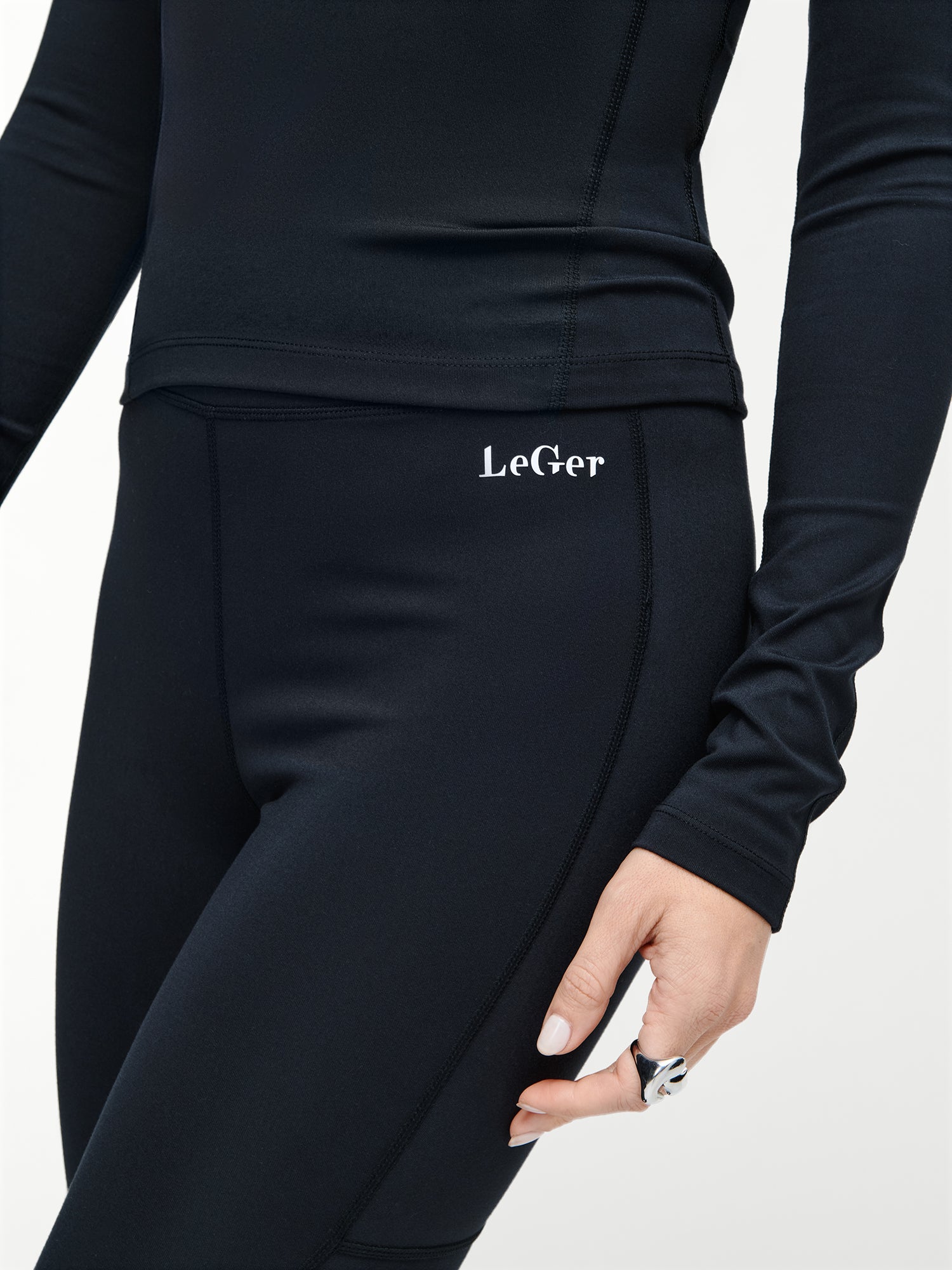 Eine Person trägt eine schwarze Thalke-Hose von LeGer by Lena Gercke mit dem Schriftzug "LeGer" am Bund und ein dazu passendes schwarzes langärmeliges Oberteil. Ihre Hand, die mit einem silbernen Ring geschmückt ist, hängt an ihrer Seite vor einem schlichten Hintergrund.