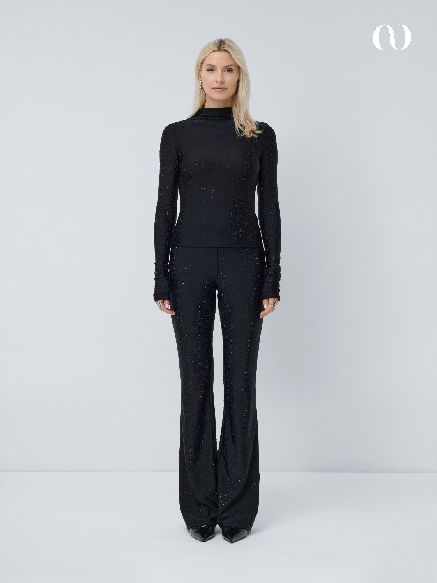 Eine Frau mit blonden Haaren blickt nach vorne und trägt das schmal geschnittene schwarze Daniela Longsleeve von LeGer by Lena Gercke und eine passende Schlaghose. Sie steht vor einem schlichten weißen Hintergrund mit einem kleinen abstrakten Logo in der oberen rechten Ecke.