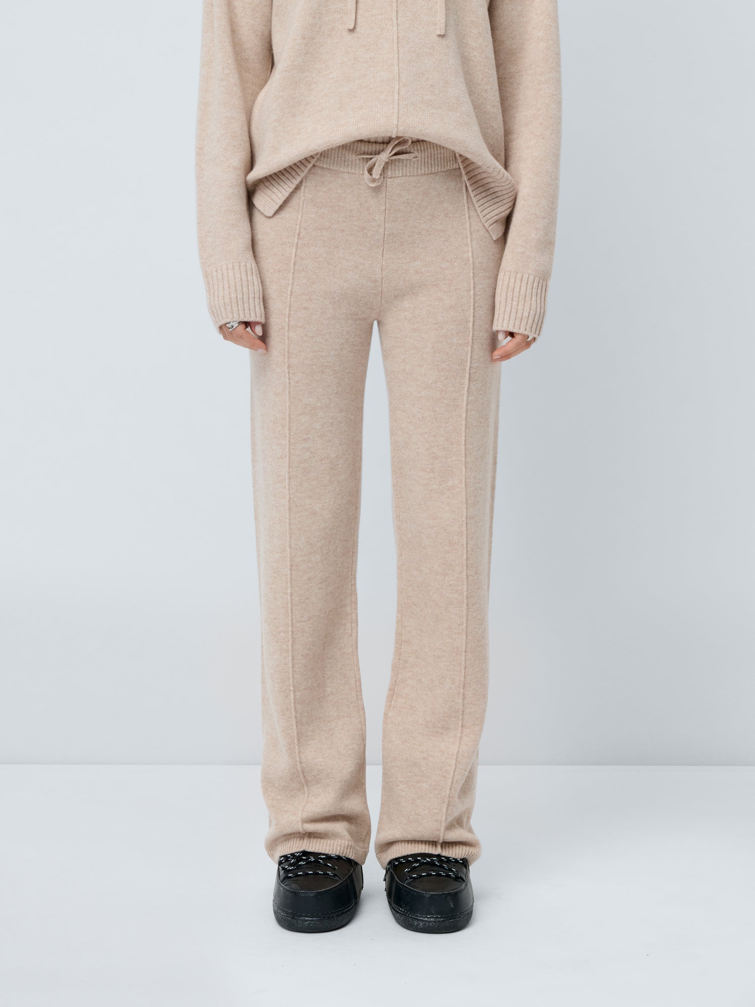 Eine Person trägt die Godja Pants von LeGer by Lena Gercke, eine beigefarbene Pull-on-Hose mit weitem Bein und lockerer Passform, gepaart mit einem passenden Pullover mit Stickerei und schwarzen Sneakers. Der Hintergrund ist einfarbig weiß; der Kopf der Person ist nicht abgebildet.