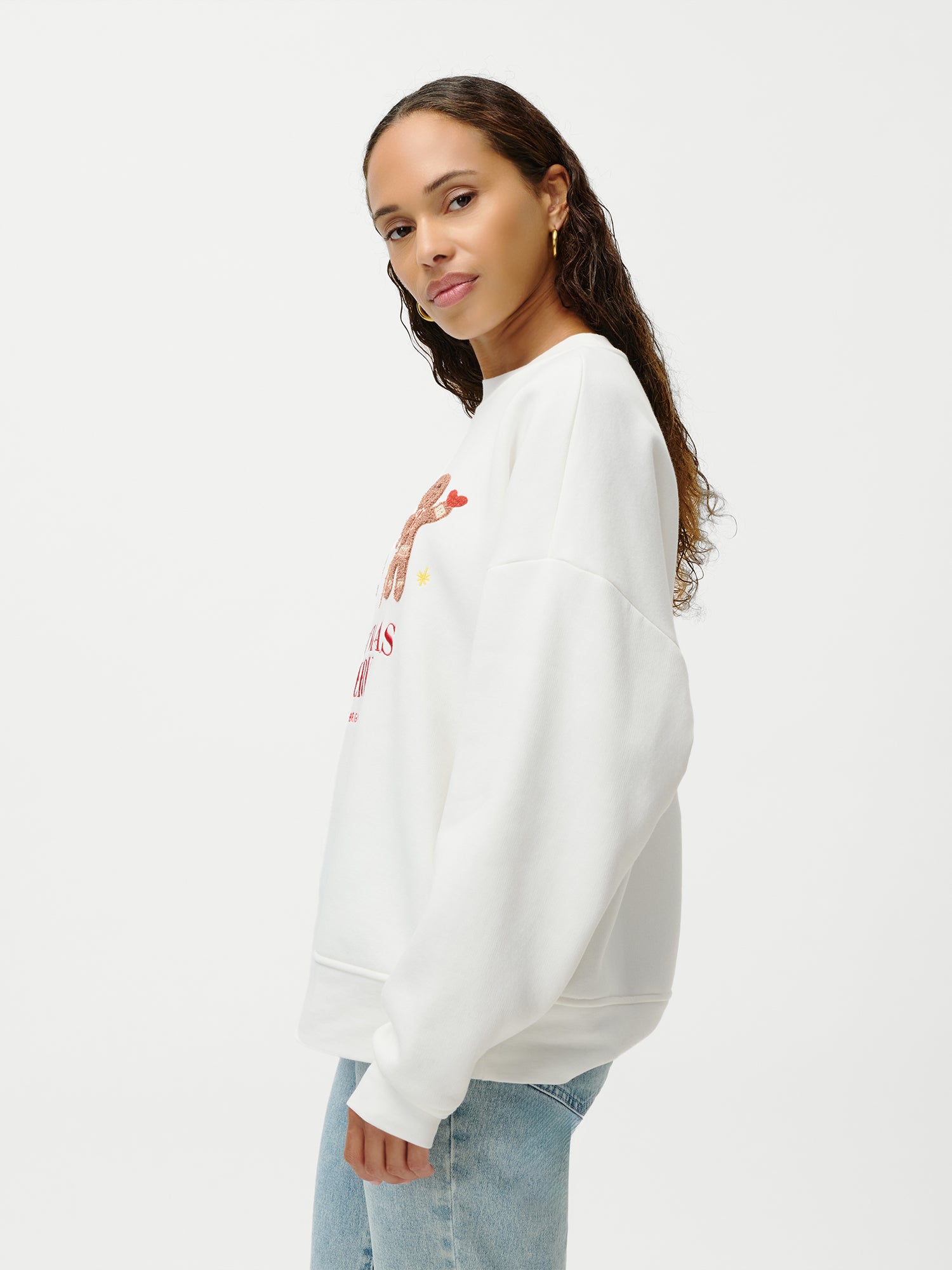 Eine Frau mit langen, lockigen Haaren trägt den Pullover „Daphne“ von LeGer by Lena Gercke und hellblaue Jeans; sie steht seitlich und blickt vor einem schlichten weißen Hintergrund in die Kamera.