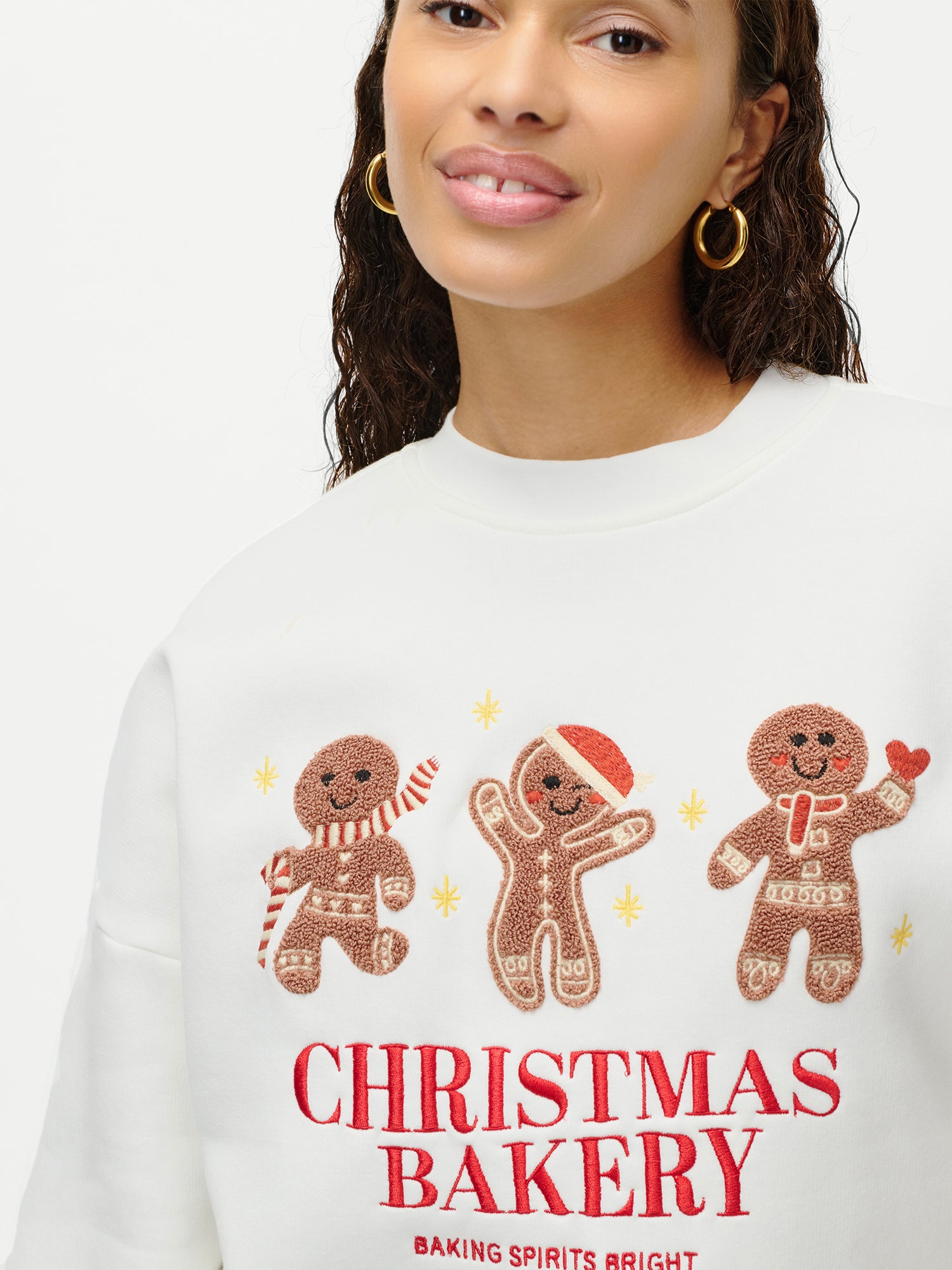 Der Pullover „Daphne“ von LeGer by Lena Gercke ist ein weißer, langärmeliger Pullover mit Rundhalsausschnitt, verziert mit gestickten Lebkuchenmännchen und dem Schriftzug „Christmas Bakery – Baking Spirits Bright“. Er wird von einer lächelnden Frau mit goldenen Creolen getragen.