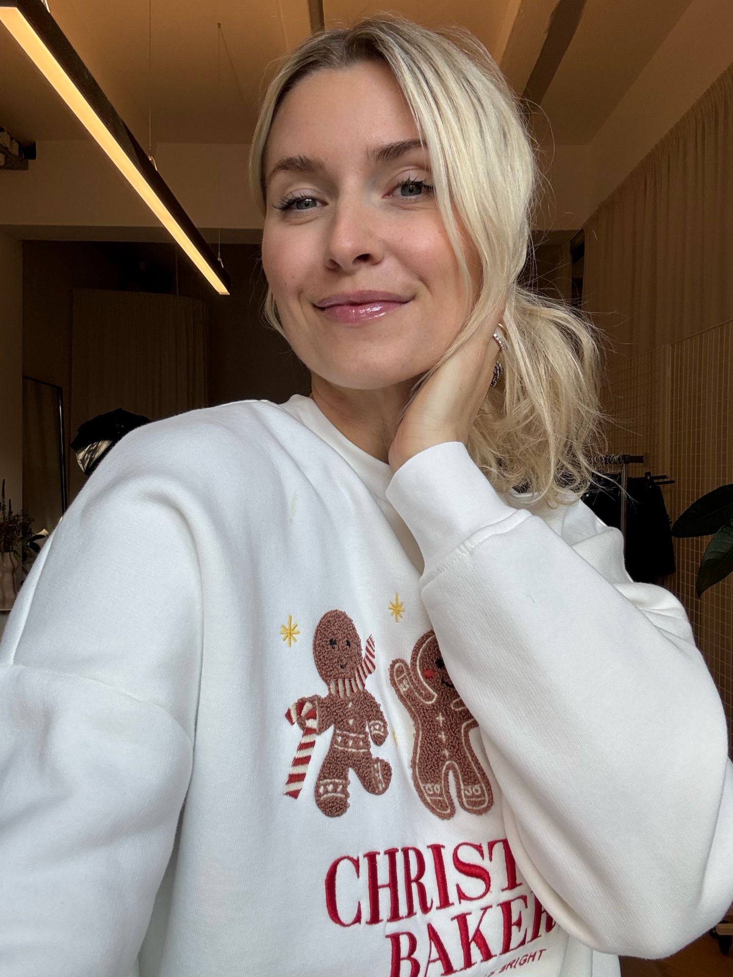 Eine blonde Frau trägt den Pullover „Daphne“ von LeGer by Lena Gercke, ein weißes Sweatshirt in normaler Länge mit Lebkuchenmännchen und der Aufschrift „CHRISTMAS BAKERY“, und lächelt sanft in einem Raum, während sie ihre Hand an den Nacken legt.
