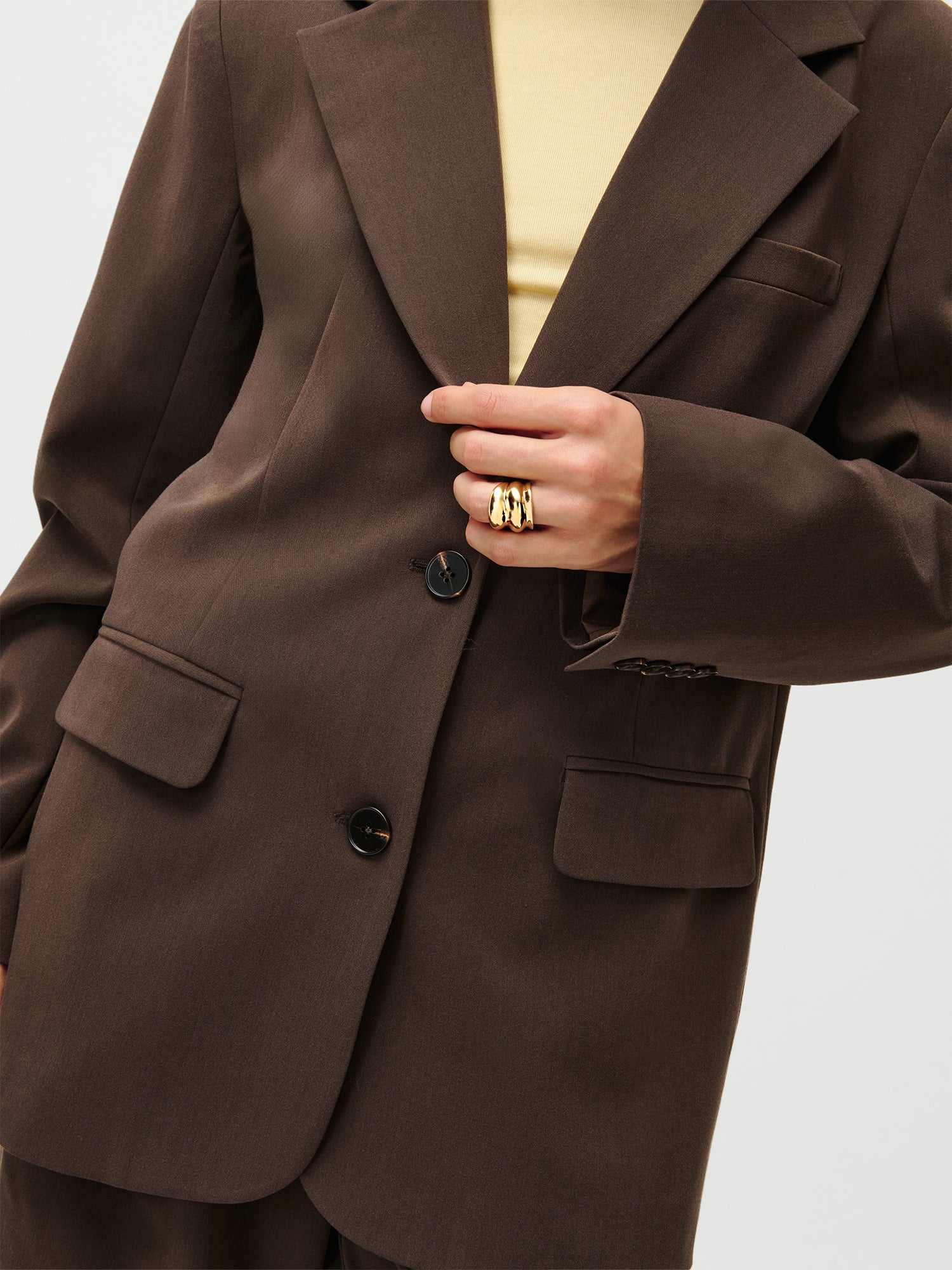 Eine Person trägt den LeGer x Purelei - Athina Blazer von LeGer by Lena Gercke in Größe 36 über einem hellgelben Top und hält das Sakko. Mehrere klobige Goldringe verzieren die Finger.