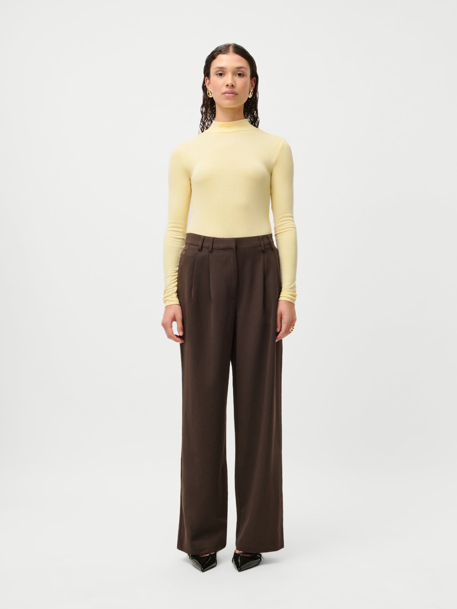 Eine Person steht mit dem Gesicht nach vorne vor einem weißen Hintergrund und trägt einen hellgelben Rollkragenpullover, die LeGer x Purelei Athina Pants von LeGer by Lena Gercke in Größe 36 und schwarze spitze Schuhe.