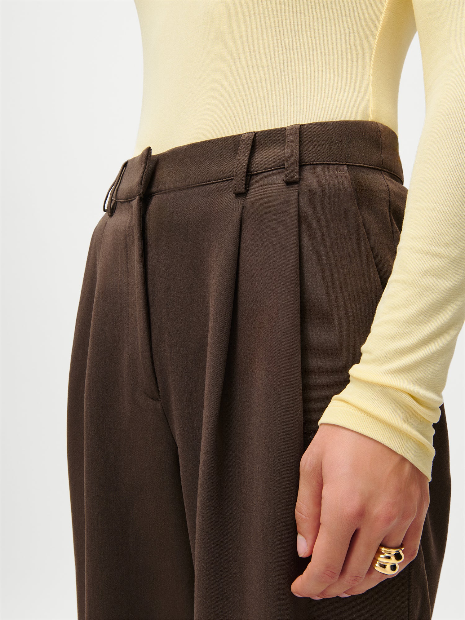 Eine Person trägt die LeGer x Purelei - Athina Pants von LeGer by Lena Gercke in Braun, gepaart mit einem taillierten, langärmeligen, hellgelben Top (Größe 36). An der Hand trägt sie einen klobigen Goldring.