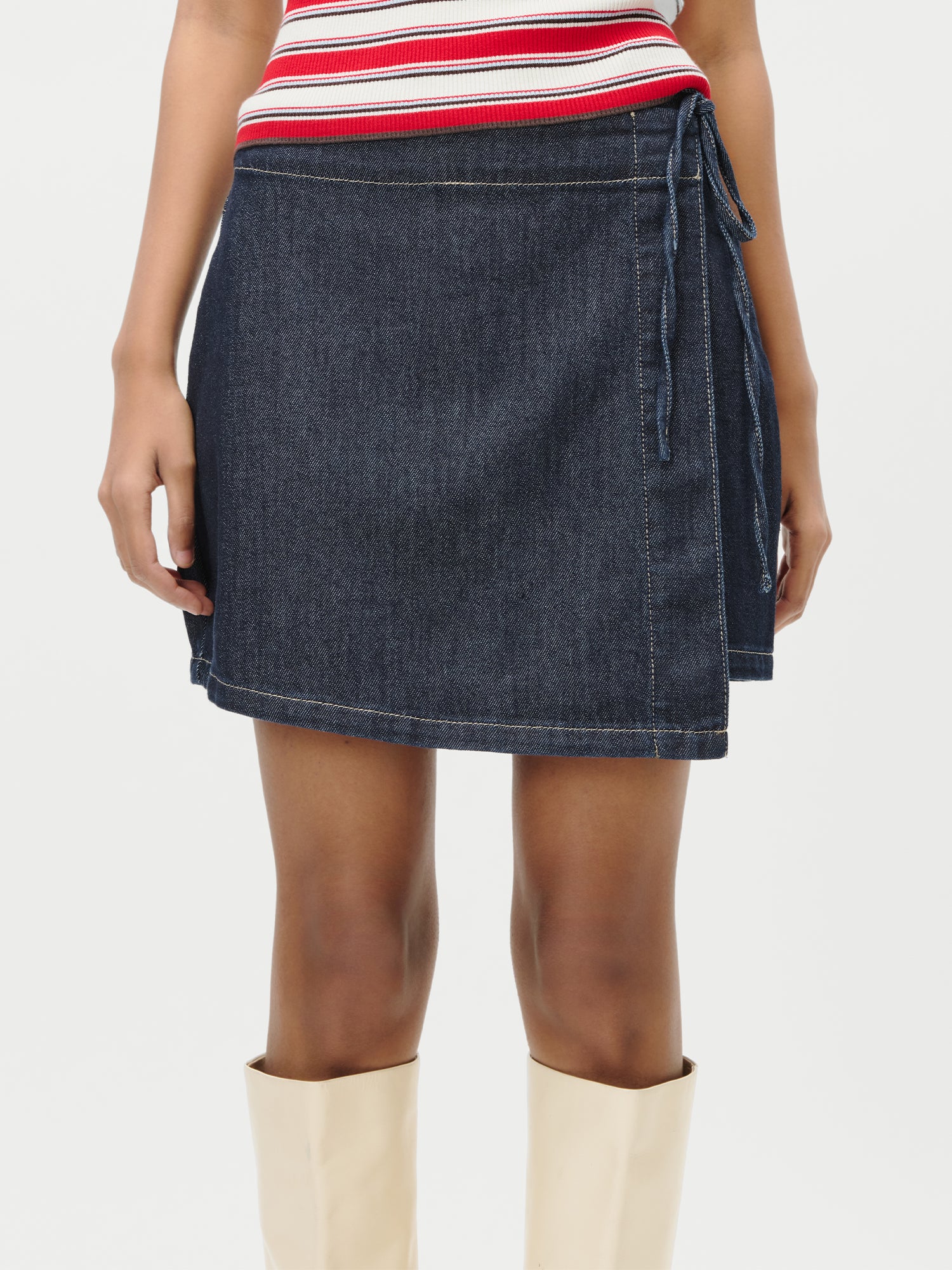 Ein Model trägt den LeGer by Lena Gercke Swantje Rock, einen hochtaillierten Wickelmini aus dunklem Denim mit seitlicher Bindung, kombiniert mit einem rot-weiß gestreiften Oberteil und cremefarbenen, kniehohen Stiefeln, dargestellt von der Taille bis zu den Knien vor einem schlichten weißen Hintergrund.