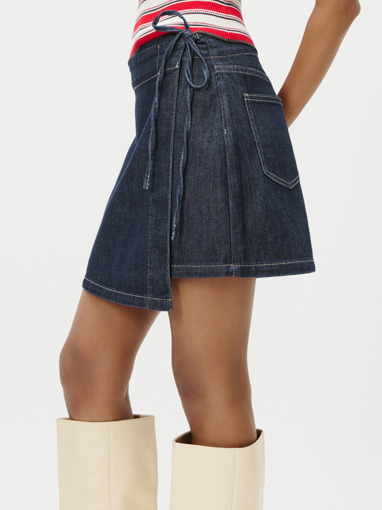 Eine Frau trägt den Wickelrock „Swantje“ von LeGer by Lena Gercke aus dunklem Denim mit hoher Taille, Seitentasche und Bindeband, kombiniert mit einem rot-weiß gestreiften Oberteil und cremefarbenen kniehohen Stiefeln vor einem einfarbigen Hintergrund.