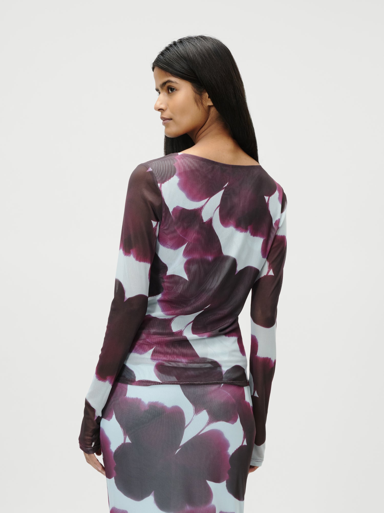 Eine Frau mit langen dunklen Haaren trägt das Floria Longsleeve von LeGer by Lena Gercke in Größe 36 mit taillierter Silhouette und großem lila-weißem Blumenmuster, während sie vor einem schlichten hellen Hintergrund über ihre Schulter schaut.