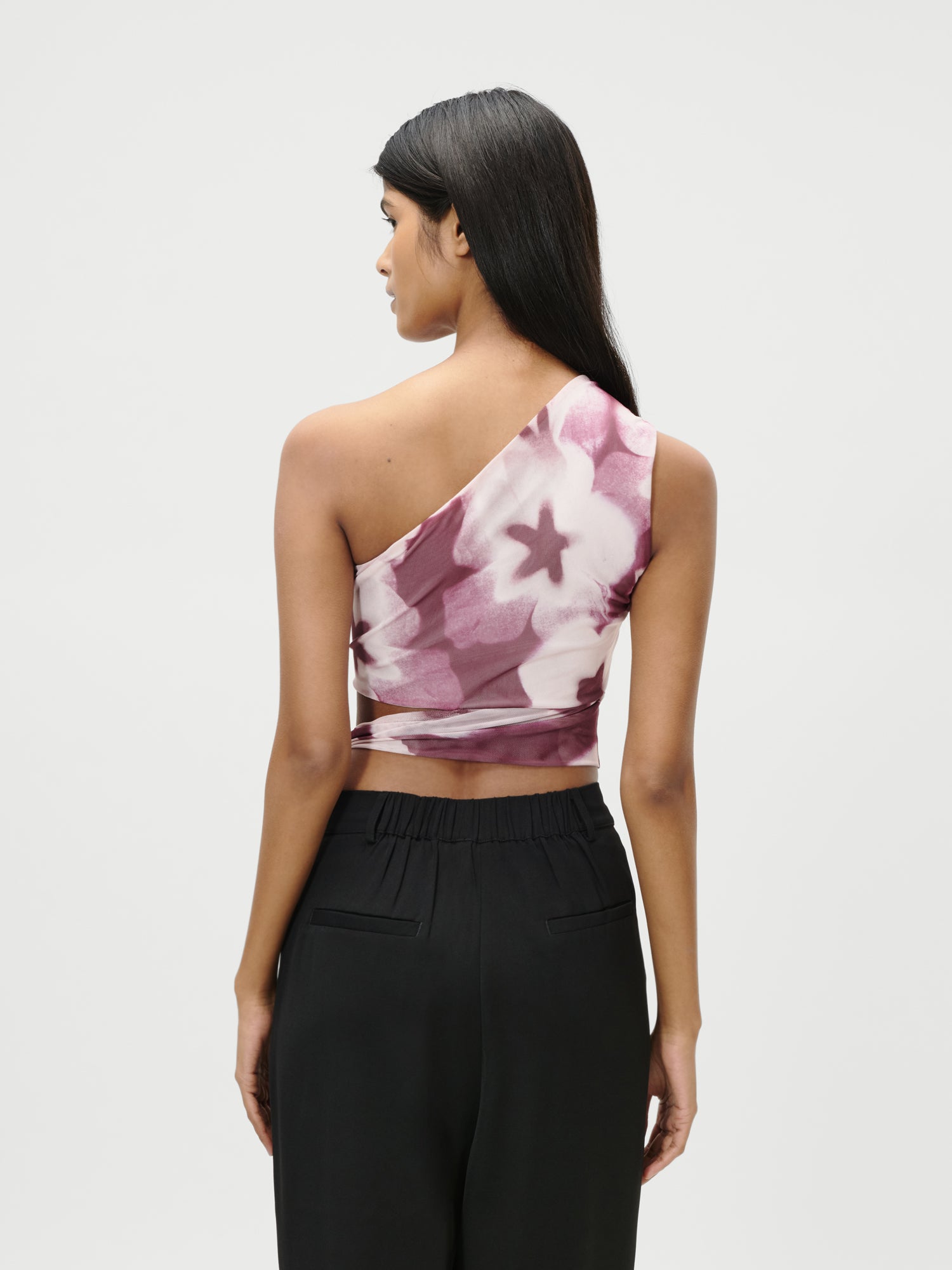 Eine Frau steht mit dem rosa-weißen, ärmellosen, geblümten Floria Top von LeGer by Lena Gercke und einer schwarzen, hochgeschlossenen Hose vor dem Hintergrund. Ihr glattes dunkles Haar fällt ihr vor einem weißen Hintergrund über den Rücken.