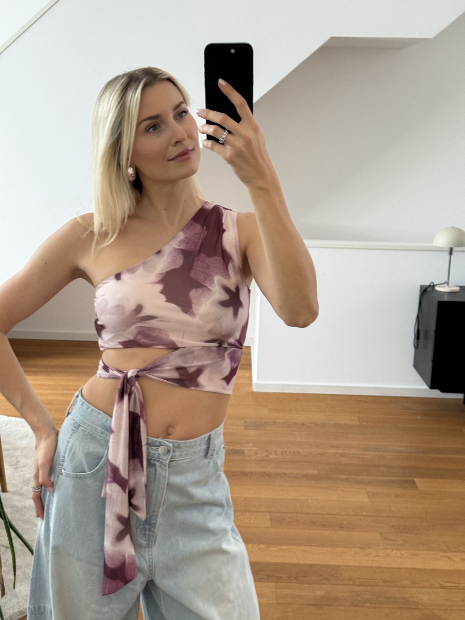 Eine blonde Frau macht ein Spiegel-Selfie in einem hellen Raum. Sie trägt das Floria Top von LeGer by Lena Gercke und hellblaue Jeans. Sie steht auf einem Holzboden mit minimalem, modernem Dekor im Hintergrund.