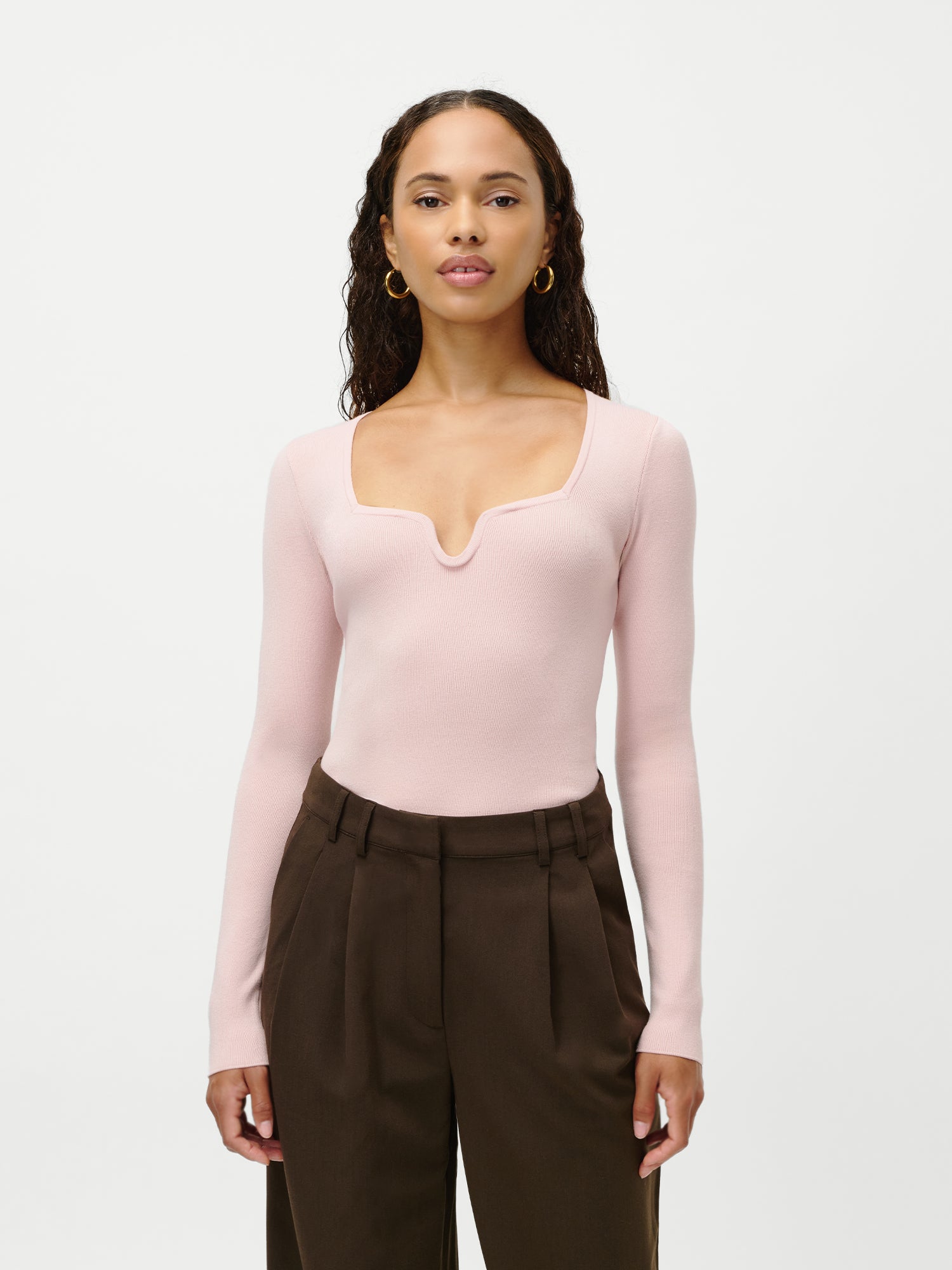 Eine Frau mit langen lockigen Haaren modelliert den LeGer by Lena Gercke Guya Jumper, ein blassrosa langärmeliges Oberteil mit viereckigem Ausschnitt, das mit einer braunen Hose mit hoher Taille kombiniert wird. Abgebildet in Größe 36 vor einem schlichten weißen Hintergrund.