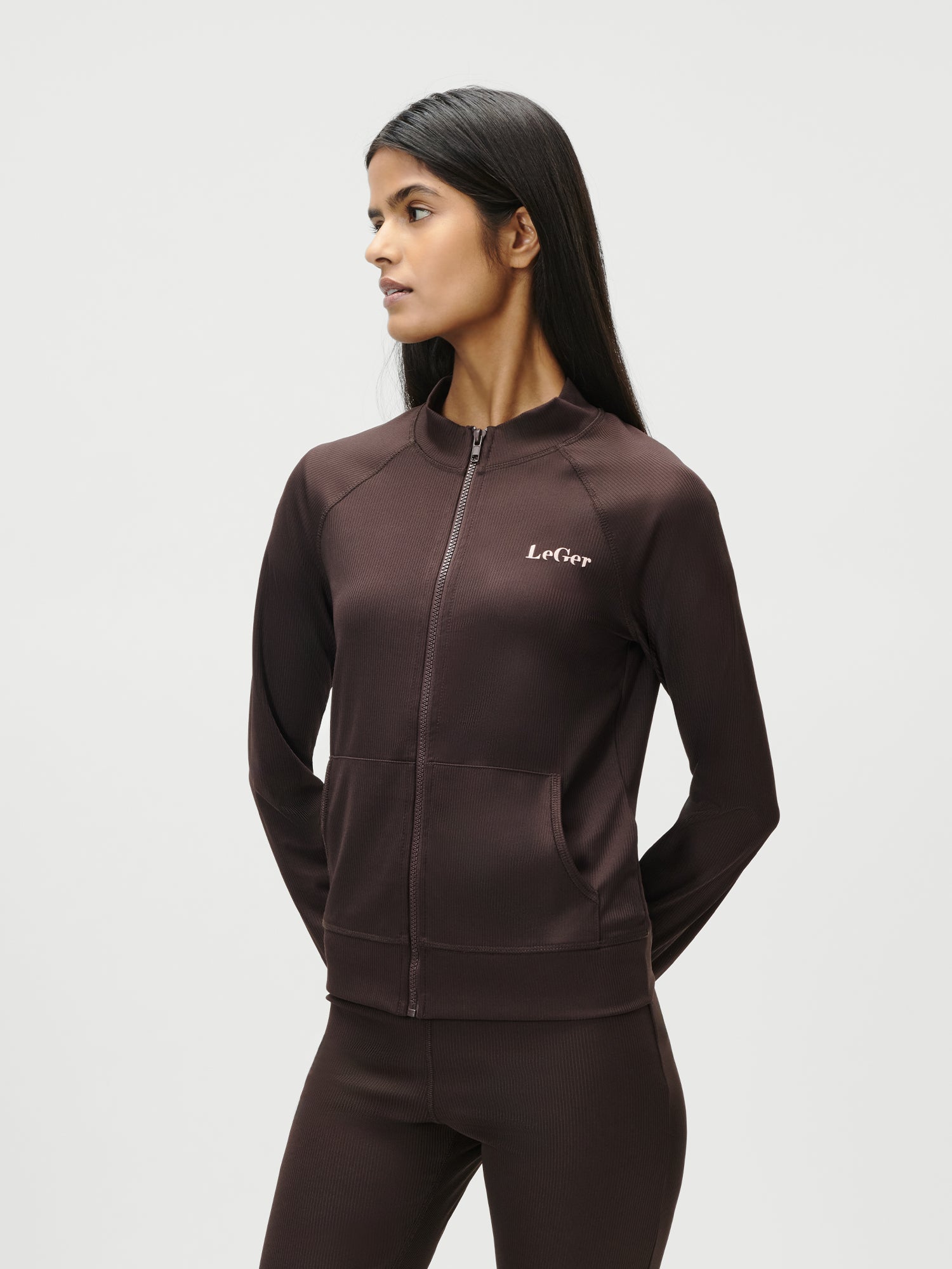 Eine Frau mit langen dunklen Haaren steht mit dem Gesicht nach links und trägt die Simone Jacket von LeGer by Lena Gercke - eine schmal geschnittene, dunkelbraune Jacke mit Reißverschluss in normaler Länge und dem Schriftzug "Le Cap" auf der Brust - sowie eine dazu passende Hose vor einem schlichten hellen Hintergrund.