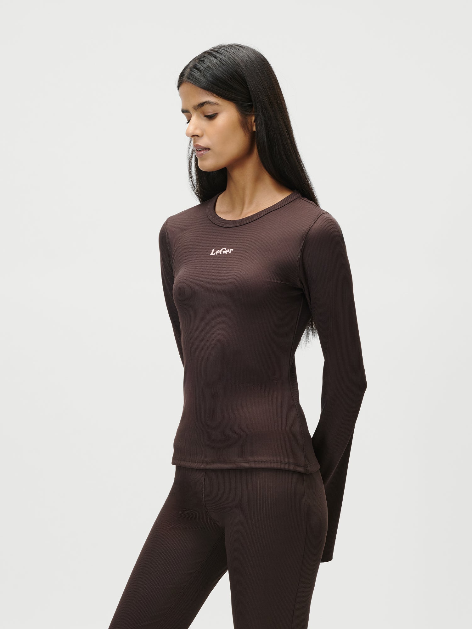 Eine Frau mit langen dunklen Haaren trägt ein tailliertes, extralanges Simone Longsleeve von LeGer by Lena Gercke und eine passende Hose in Größe 36. Das braune Oberteil ist mit einem kleinen weißen Logo auf der Brust versehen. Sie steht vor einem schlichten weißen Hintergrund und blickt mit entspannten Armen nach unten.