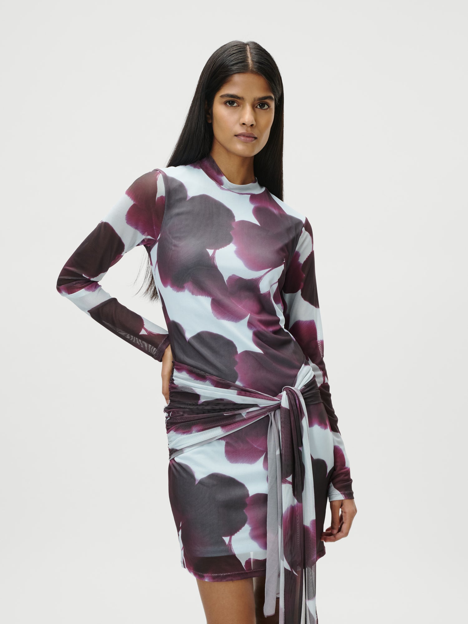 Eine Frau mit langen dunklen Haaren trägt das Albany Dress von LeGer by Lena Gercke, ein figurbetontes, langärmeliges Kleid mit lila-weißem Blumenmuster und Bindeband in der Taille, vor einem schlichten weißen Hintergrund.