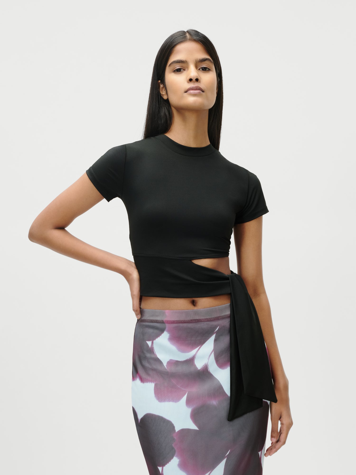 Eine Frau trägt das Leia T-Shirt von LeGer by Lena Gercke mit figurbetontem Schnitt, kurzem Arm und Taillen-Bindeband, kombiniert mit einem lila Blumenrock, vor schlichtem Hintergrund.