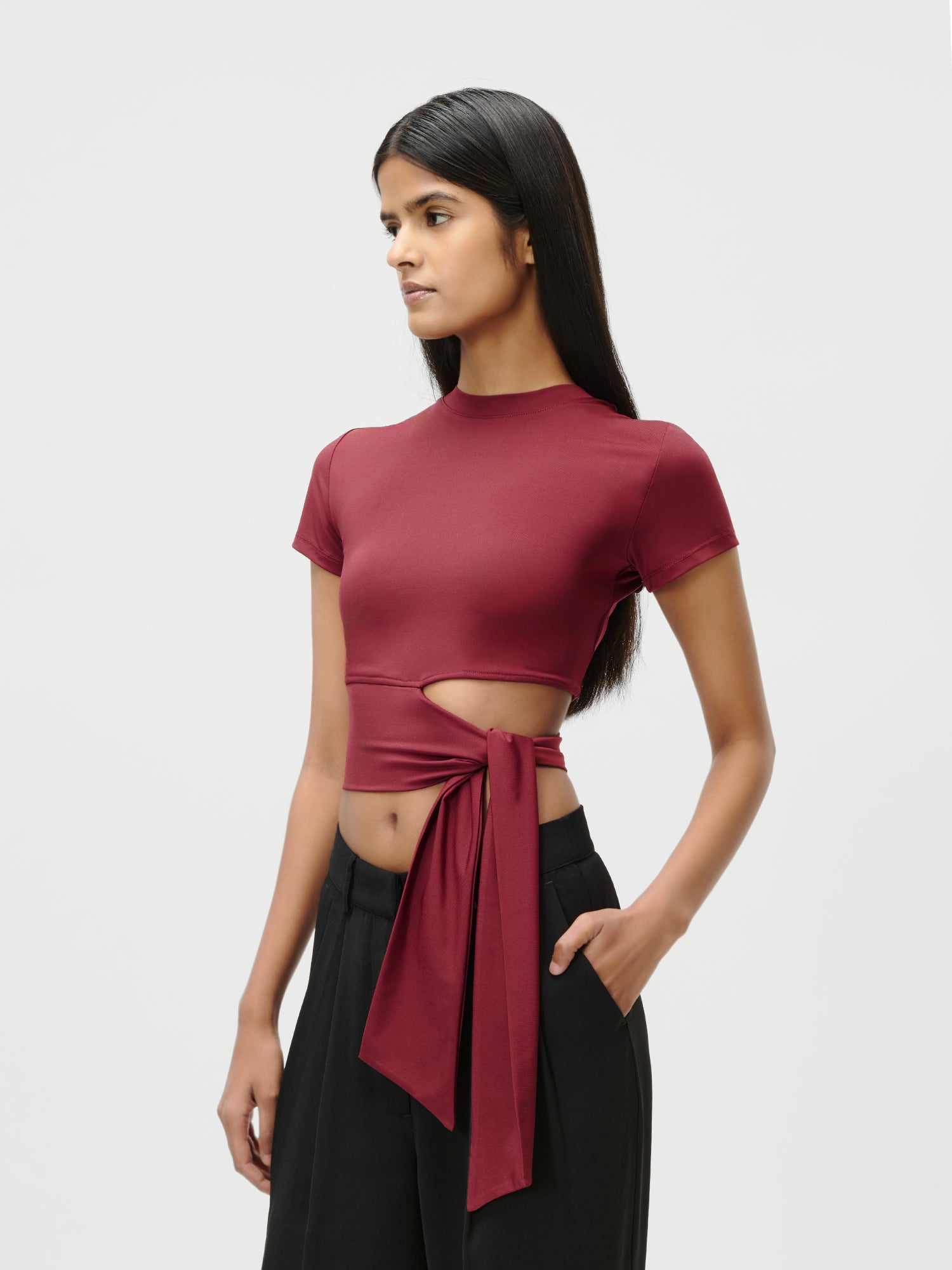 Eine Frau mit langen dunklen Haaren trägt das burgunderfarbene Leia T-Shirt von LeGer by Lena Gercke. Es ist tailliert geschnitten, hat kurze Ärmel, einen seitlichen Ausschnitt und eine große gebundene Schleife. Sie kombiniert es mit einer schwarzen Hose mit hoher Taille und steht vor einem schlichten, hellen Hintergrund.
