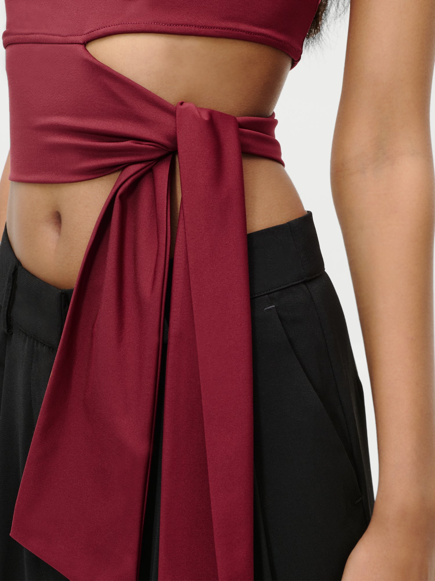 Eine Nahaufnahme zeigt eine Person, die das burgunderfarbene Leia T-Shirt von LeGer by Lena Gercke trägt, das mit einem figurbetonten Wickelschnitt und einer Fliege in der Taille versehen ist, gepaart mit einer schwarzen Hose mit hoher Taille. Das Bild hebt die Details des Knotens und des Stoffes hervor.