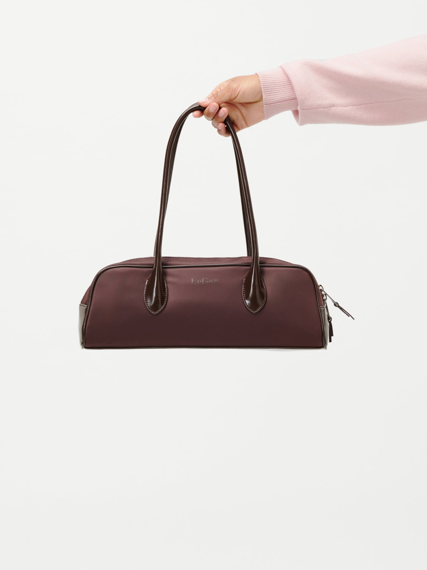 Eine Person in einem hellrosa Ärmel hält die LeGer by Lena Gercke Frauke Bag, eine kastanienbraune Handtasche mit dunkelbraunen Griffen und Verzierungen, vor einem schlichten weißen Hintergrund.