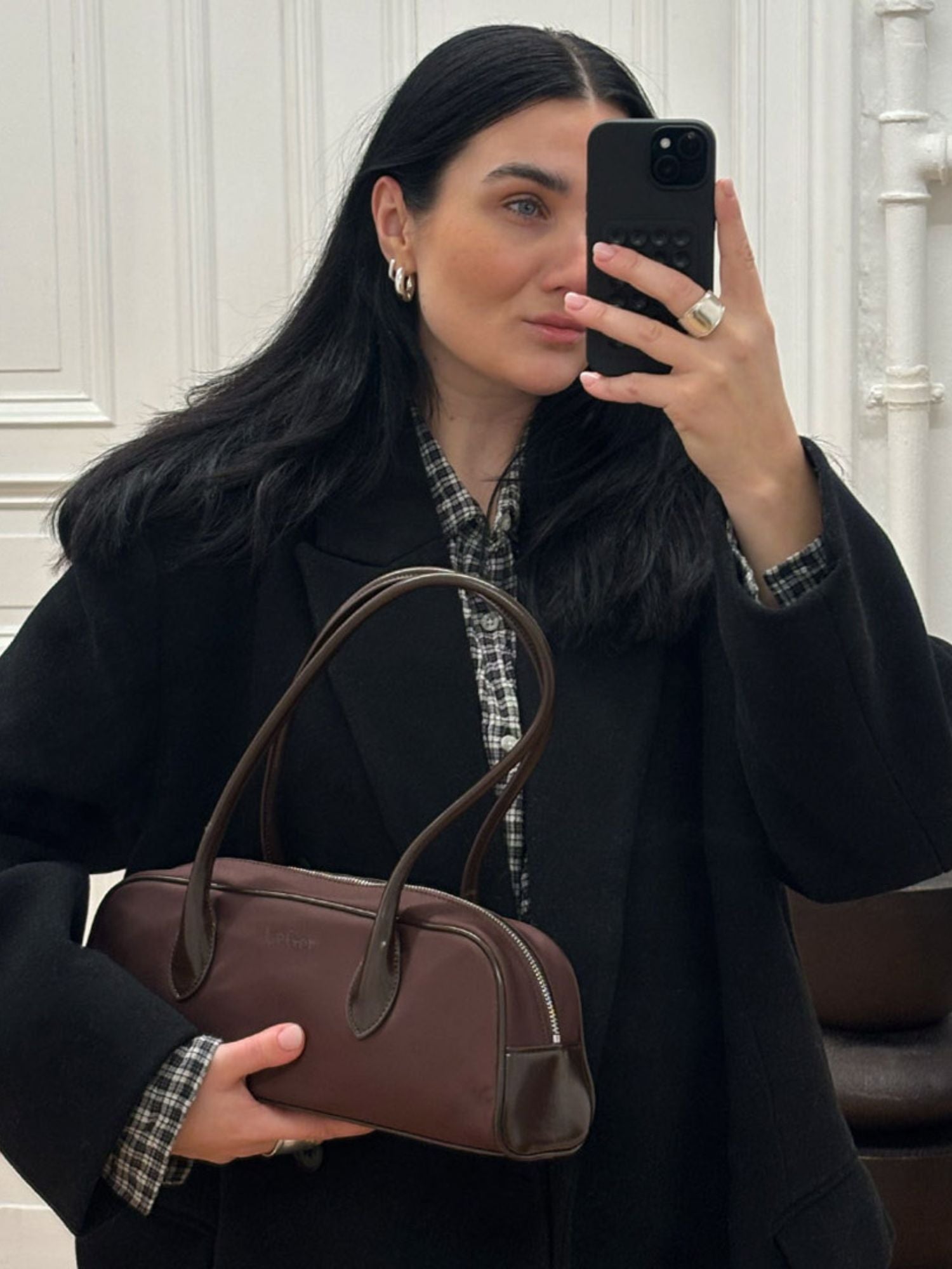 Eine Frau mit langen dunklen Haaren macht ein Selfie im Spiegel, trägt einen schwarzen Mantel über einem karierten Hemd und hält die Frauke Bag von LeGer by Lena Gercke in der Hand. Sie trägt dazu Ohrringe und goldene Ringe und posiert vor einer weiß getäfelten Wand.